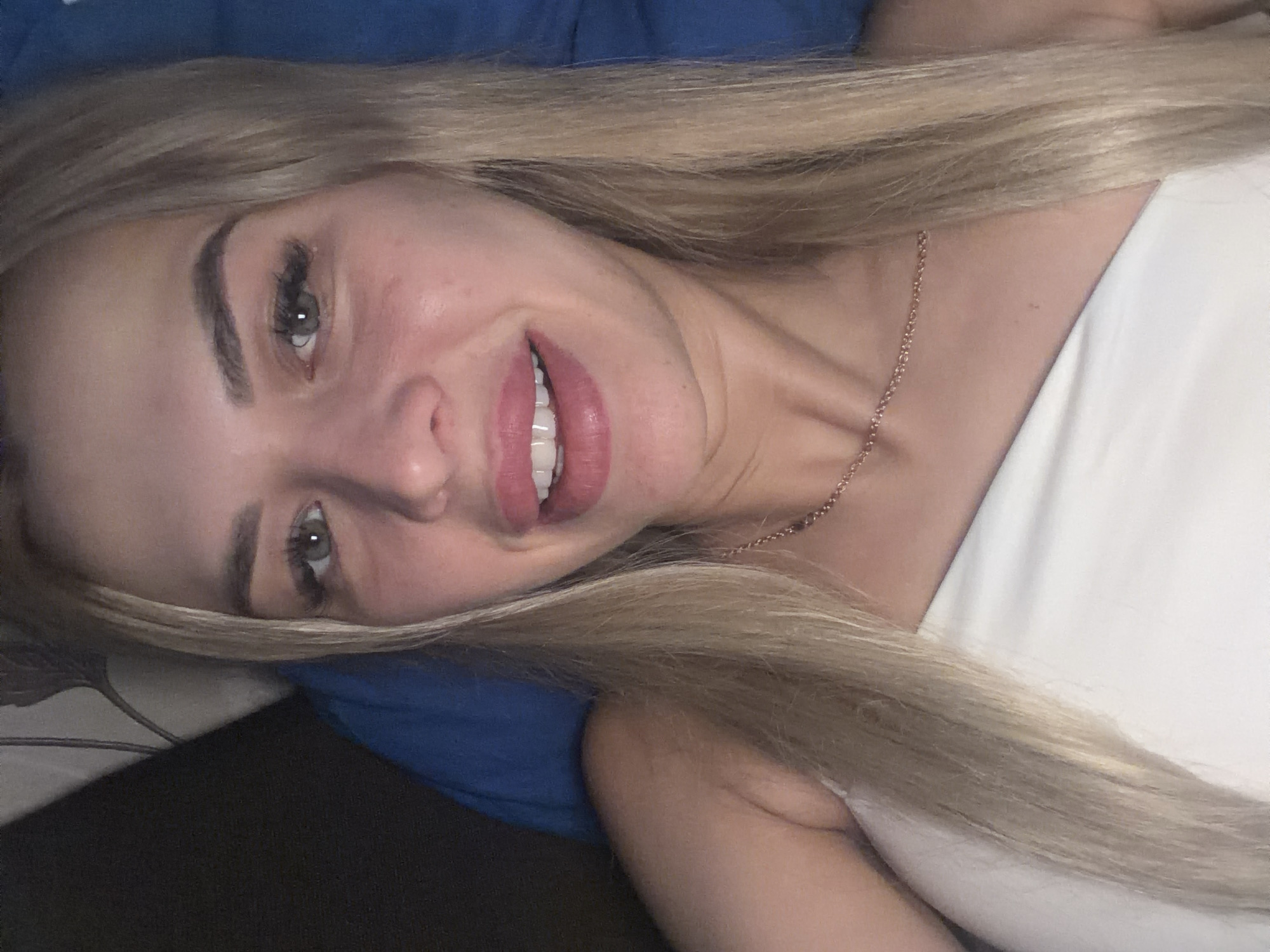 This is a close-up, high-angle selfie of a young Caucasian woman with blonde hair and light-colored eyes. She is wearing a white off-the-shoulder top and a delicate gold necklace. Her lips are full and painted with a glossy pink lipstick, and she is smiling. The background appears to be a bed with blue and patterned bedding. The lighting suggests an indoor setting, likely at night or in a dimly lit room. There is no indication of the location being in Kyiv, Ukraine.