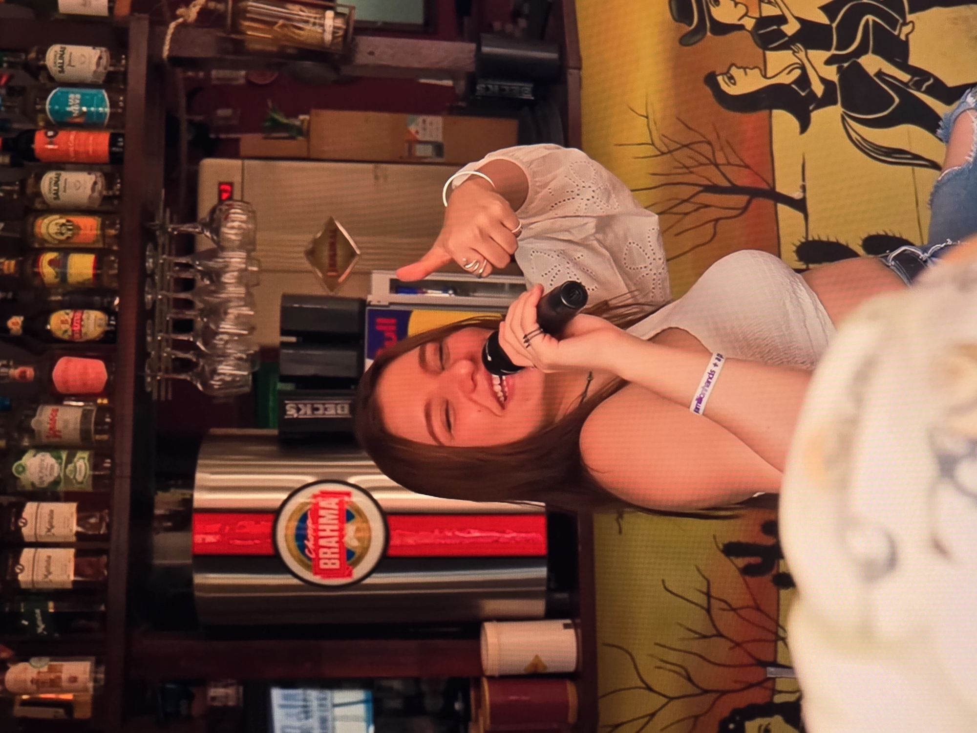 A young woman with long brown hair and a white crop top is holding a microphone and singing. She is smiling and giving a thumbs-up with her right hand, which is adorned with several silver rings and a bracelet. She is wearing blue jeans. Behind her, a Brahma beer dispenser is visible. The background shows a shelf with various bottles, wine glasses hanging, and a mural depicting a desert scene with cacti and people riding horses. The overall setting appears to be a bar or restaurant in Natal, Brazil, suggested by the presence of the Brahma brand, a popular Brazilian beer.