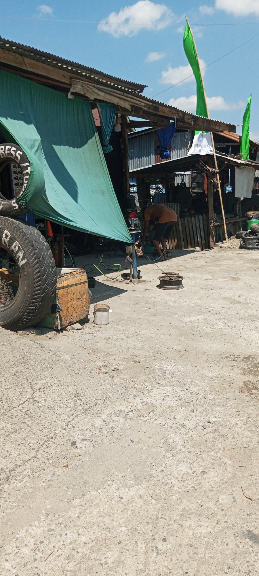 This is a photograph taken on a sunny day in Lempa, Indonesia, depicting an outdoor workshop. In the foreground, a concrete surface shows signs of wear and tear, with some cracks and embedded stones. To the left, a large tire is prominently displayed, with white lettering partially visible. Next to the tire is a rust-colored metal object, possibly a container. A grey metal bucket is also present.
In the middle ground, a man, wearing a brown shirt and dark shorts, is bending over a piece of machinery. He appears to be working on it, with tools and equipment around him. A bright green tarp is stretched out over a section of the workshop, providing shade. Various other objects, including what looks like scrap metal and possibly spare tires, are scattered around the area. In the background, a rustic structure with a corrugated metal roof is visible, suggesting a simple building or stall. Green flags are flying from poles above the structure. The overall impression is that of a busy, functional workshop in a somewhat informal setting.