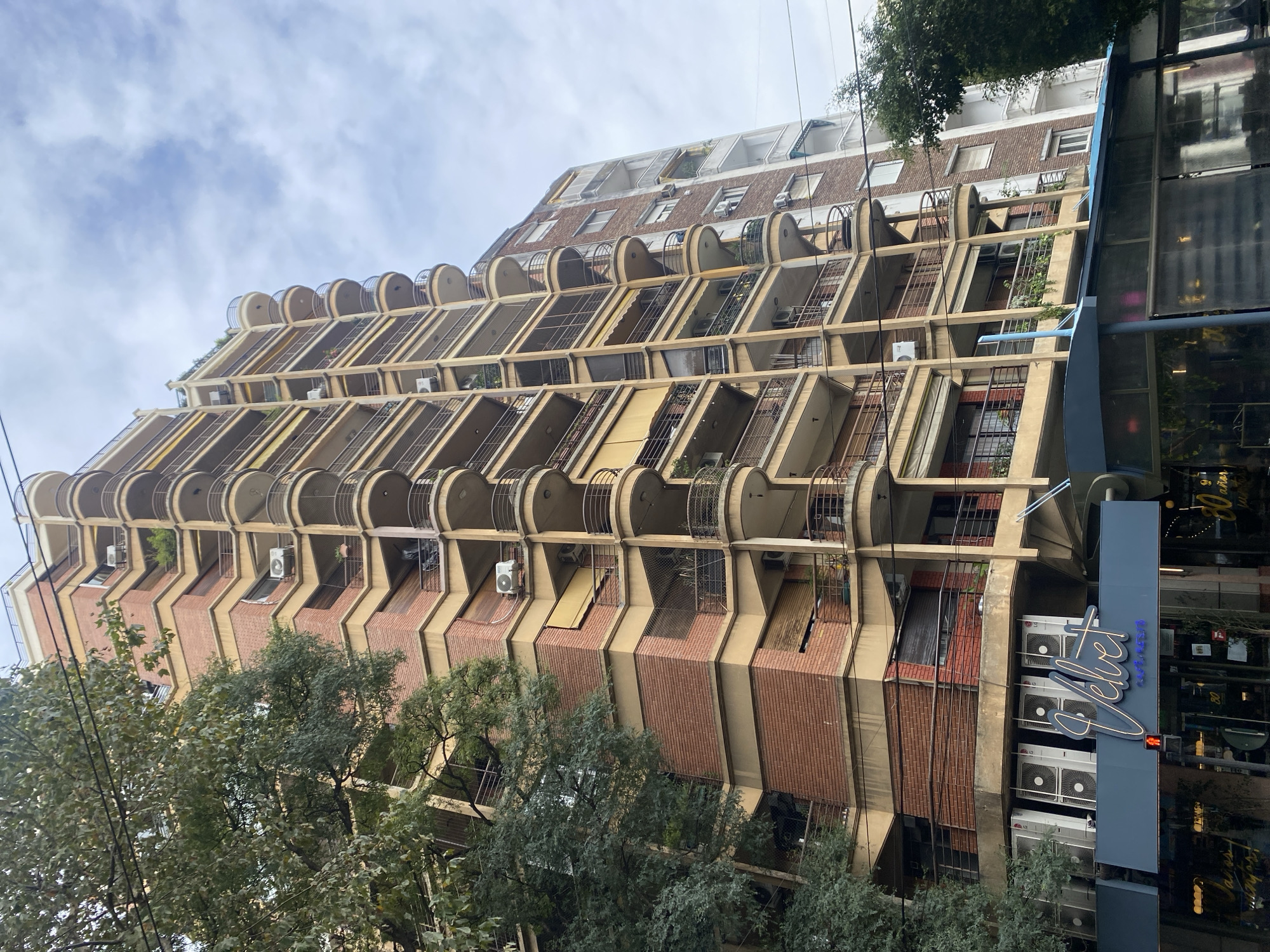 An upward-angled, daytime photograph captures a distinctive multi-story building in Buenos Aires, Argentina, under a bright, cloudy sky. The prominent structure features a unique architectural design with tiered, recessed balconies that step back as the building rises.
The building's facade is a mix of light-colored concrete or stucco and red brick paneling. The upper floors are characterized by numerous semicircular or cylindrical enclosed balconies with metal railings, while the lower and middle floors exhibit deep, rectangular, recessed balconies, many faced with red brick. Air conditioning units are visible on several balconies, and some greenery suggests potted plants.
To the right, at the ground level, a commercial establishment with large glass windows is partially visible. A prominent blue sign with white neon lettering spells out "Velvet," with additional, smaller text below it that appears to read "pastelería." Reflections in the glass show interior details and distorted shapes.
In the foreground and along the left side, dense green trees partially obscure the lower and middle sections of the building. The sky is largely overcast with soft, scattered clouds. No discernible human activity or specific event is taking place, presenting a static scene of urban architecture.