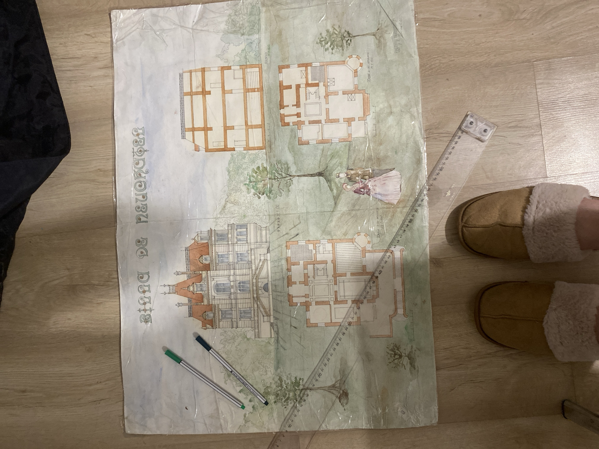 The image depicts a floor-level photograph of an architectural drawing on a light brown wooden floor. The drawing is rendered in watercolor and ink, and it appears to be a depiction of a house or estate. The name "Birra De naueRGEL" is written along the left side of the drawing in a vertical orientation. A transparent plastic ruler with metric measurements rests diagonally across the drawing, from top right to bottom left. A pair of pens, one green and one dark blue, lie near the bottom left corner of the drawing. In the upper left corner, a portion of a black, possibly velvet, garment is visible. To the right of the drawing, a pair of brown slippers with white fur lining are present, with a leg visible inside one of the slippers. The drawing includes architectural plans labeled "Pleae M 1:00", "Тилан 1-10 атка M-1100", and "План разка". The drawing contains details of the buildings and a landscape with figures. There is no immediate visual indication of the city of Yekaterinburg, Russia in the image.