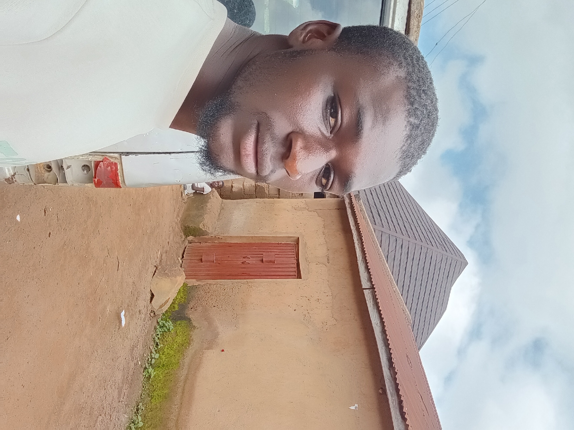 A selfie is taken from the chest up of a Black male with short, dark hair and a goatee, looking towards the camera. He is wearing a light-colored t-shirt. The background suggests a rural or semi-urban setting in Bukuru, Nigeria. To the left of the man is the side of a white vehicle. Behind him, a light brown building with a red corrugated metal roof is visible. This building has a red wooden door and appears to be constructed of mud or stucco. Further back, a darker, pyramid-shaped roof is visible. A small child in a white shirt can be seen standing near the building on the right. The ground is dirt, with some patches of green vegetation. The sky above is partly cloudy.