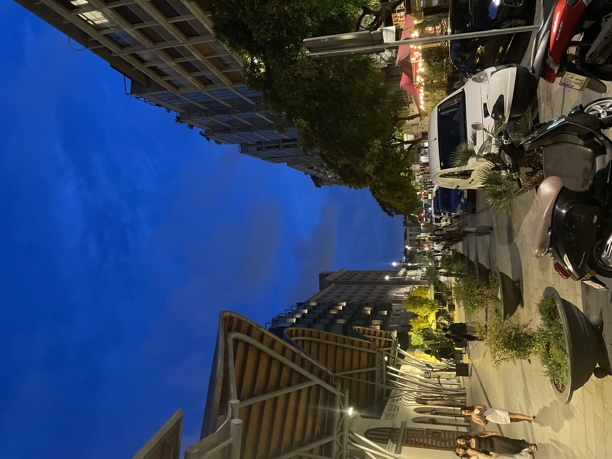 This image depicts a nighttime street scene in Barcelona, Spain, specifically along the perimeter of the Mercat de Sant Antoni (Sant Antoni Market). The sky is a deep indigo blue with subtle cloud formations.
On the left, the distinctive, undulating roof structure of the Mercat de Sant Antoni dominates the upper-left quadrant. This structure features a light-colored metal framework and a ribbed, light-brown underside, likely wood or a similar material. Below it, the market building is white with arched openings and windows. Several light fixtures illuminate the underside of the market's roof. Further back on the left, a modern building with numerous windows and balconies is visible.
The center and right side of the image show a wide urban street flanked by buildings, trees, and street furniture. A row of mature trees with dense green foliage lines the sidewalk on the right, some illuminated by artificial lights. Behind these trees, multi-story buildings with balconies and illuminated windows are visible. Further down the street on the right, red awnings suggest outdoor dining areas, with string lights visible beneath them.
The wide sidewalk in the foreground and midground is paved with light-colored flagstones. Dark grey, flared planters containing various green plants are spaced along the sidewalk. Several individuals are present:
* In the lower-left, two women walk towards the viewer; one in a long, dark, sleeveless dress and the other in a short, white, sleeveless dress, carrying a small bag.
* Further back on the sidewalk, other pedestrians are visible, some indistinct due to distance and lighting.
On the street, vehicles are parked and present. A prominent white commercial van (likely a Citroën Berlingo or Peugeot Partner variant) is parked facing the viewer in the midground, positioned near the planters. In the immediate foreground, a dark scooter with a light brown seat and a red scooter are parked, partially obscured by the planters. Cars are visible parked and potentially moving further down the street in the background. Streetlights cast artificial illumination across the scene, contrasting with the dark sky.
