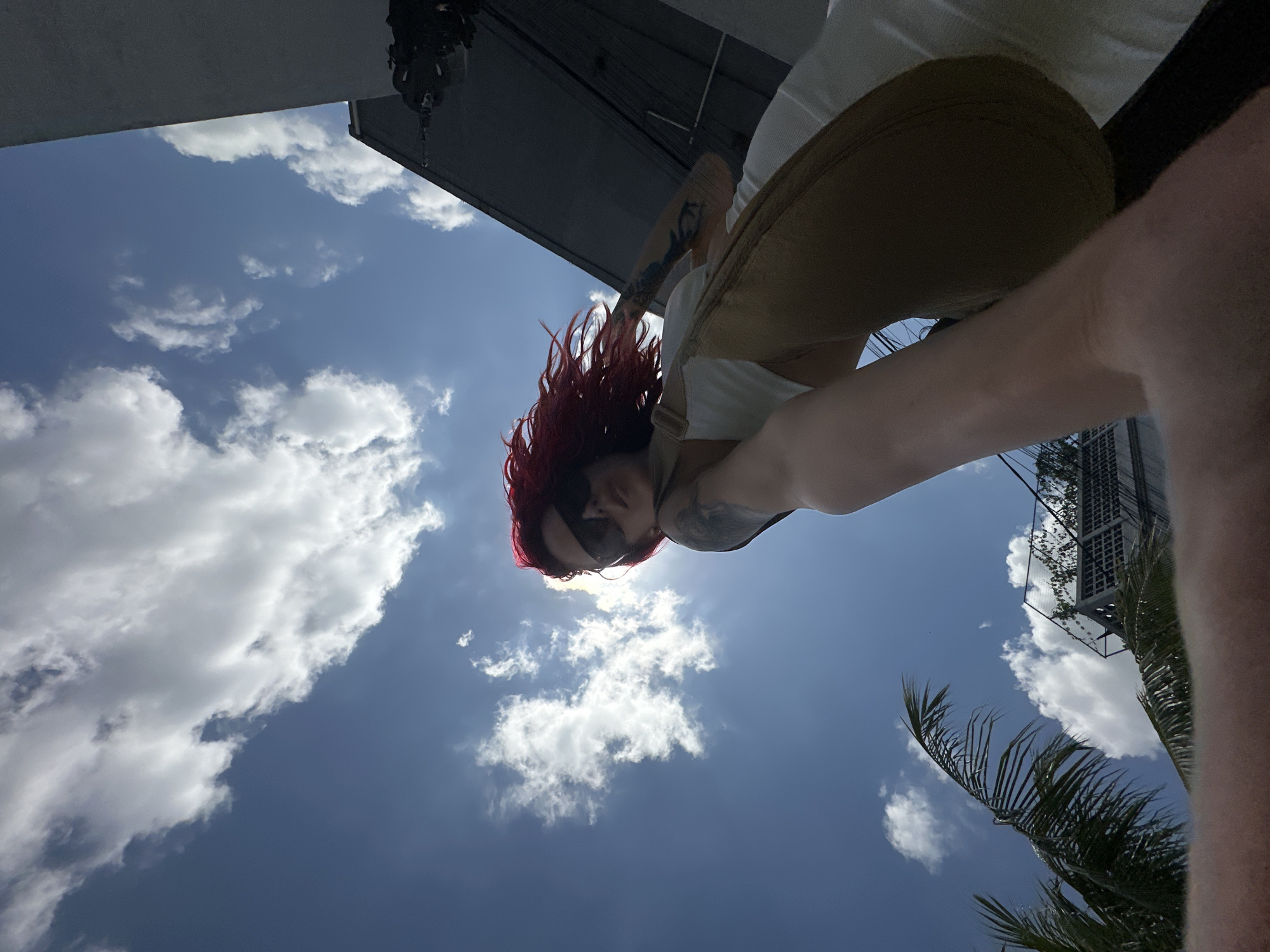 In North Kuta, Indonesia, a low-angle, wide shot captures a person with vibrant red hair and sunglasses against a bright blue sky filled with fluffy white clouds. The person is wearing a white top and tan overalls, and their arm extends towards the camera. In the background, a palm frond is visible on the left, and on the right, a modern building structure with a grid pattern and some greenery can be seen, suggesting an urban or resort setting. The sun is positioned behind the person, creating a lens flare effect and casting a bright glow.