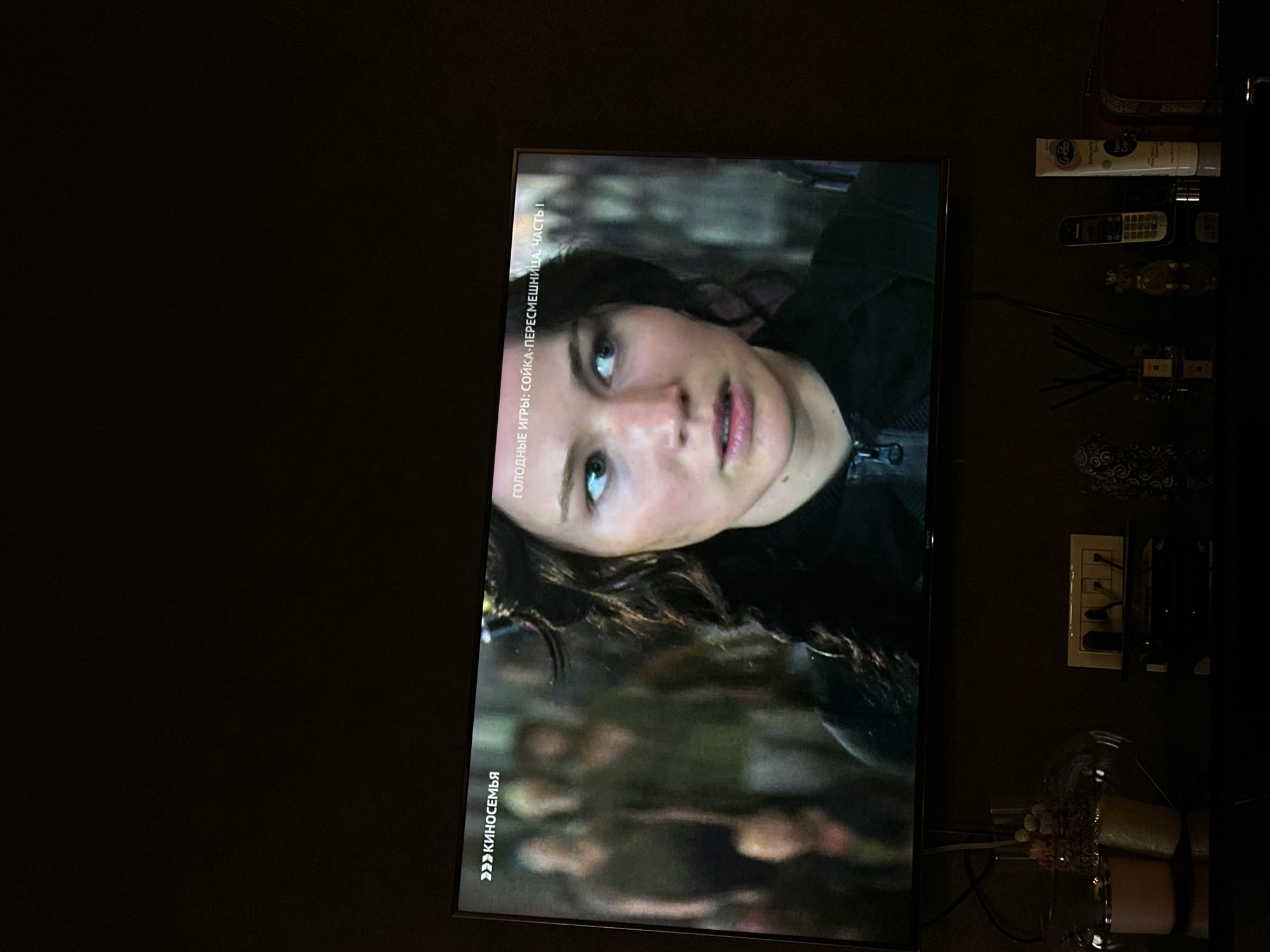 The image shows a television screen displaying a scene from the movie "The Hunger Games: Mockingjay - Part 1." On the screen, actress Jennifer Lawrence is visible. To the right of the television, various household objects are arranged on a dark surface. These include a tube of "G-PAW" hand cream, a remote control, a decorative figure, an air freshener with diffuser sticks, and a decorative dish containing candles. The scene appears to be indoors, with a dark background suggesting low lighting in the room. There is no visual indication of the location being Yerevan, Armenia.
