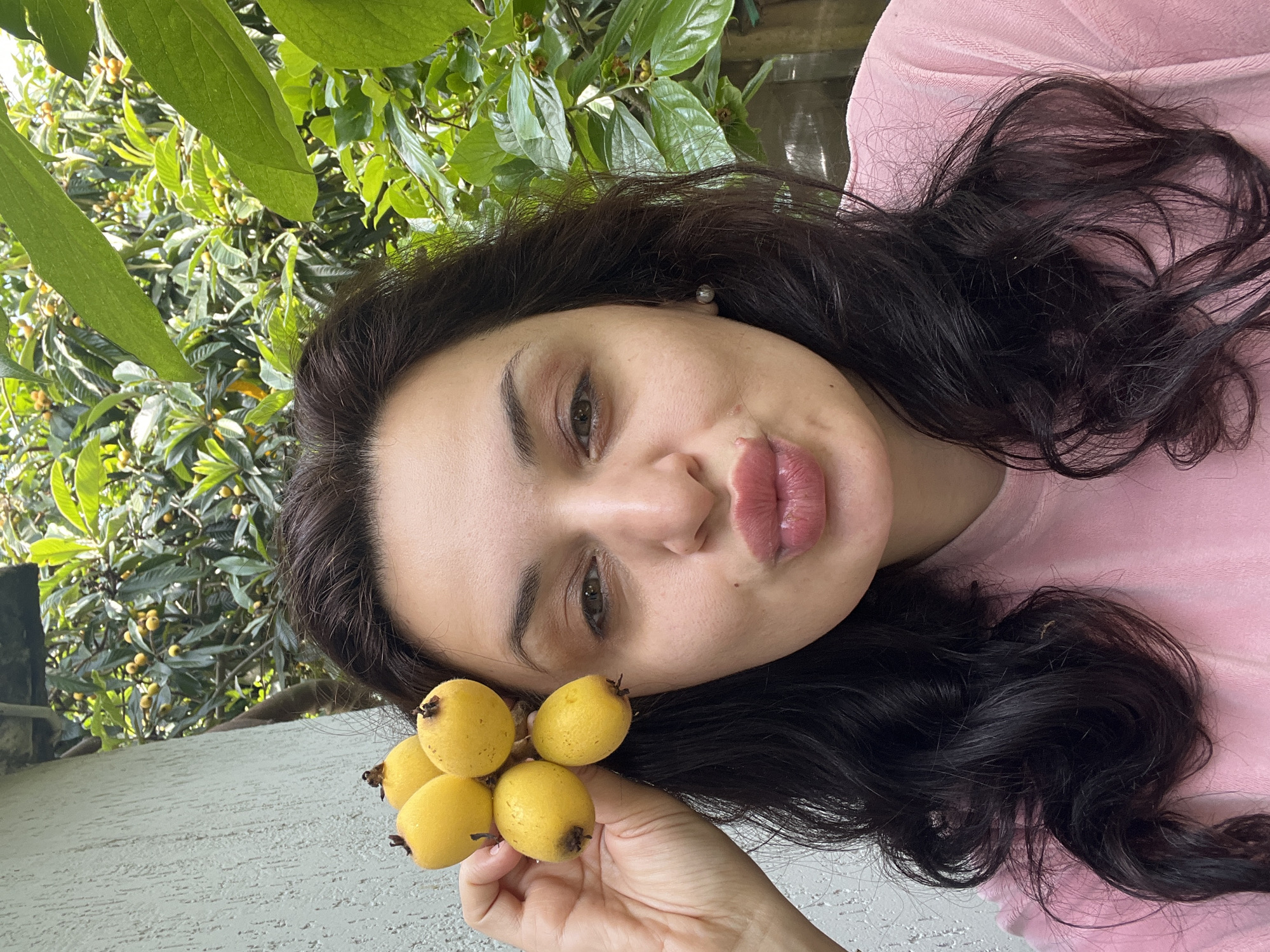 A medium close-up, outdoor selfie captures a woman holding a cluster of five small, oval-shaped, yellow fruits, likely loquats, in her left hand near her temple. She has dark, wavy hair and fair skin. Her eyes are open and directed towards the camera, while her lips are pursed in a "kissing face" expression. She wears a pink, textured top and a light-colored stud earring is visible in her right ear. The background on the left features a light grey, textured wall. To the right and top, dense green foliage from a tree is visible, with multiple small yellow fruits scattered among the leaves, mirroring the fruits she holds. The scene is brightly lit by natural light in an outdoor setting in Unknown, Unknown.