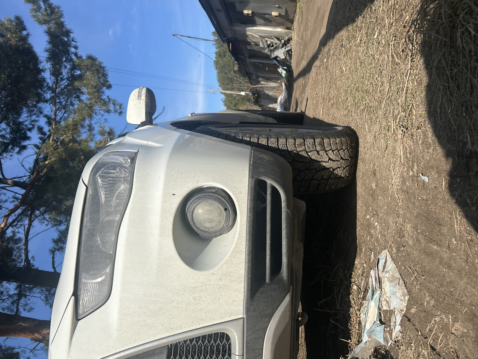 The image shows a close-up of the front end of a dirty white vehicle parked on a dirt road. The left side of the frame is dominated by the vehicle's headlight, fog light, and a portion of the grille. A tree and blue sky are visible above the car. To the right of the car is the dirt road, a tire of the vehicle, some trash and a row of garage structures. In the background, trees appear to line the horizon. While the location of the scene is Chaikovsky, Russia, the image does not have enough content to determine the exact location within the city.