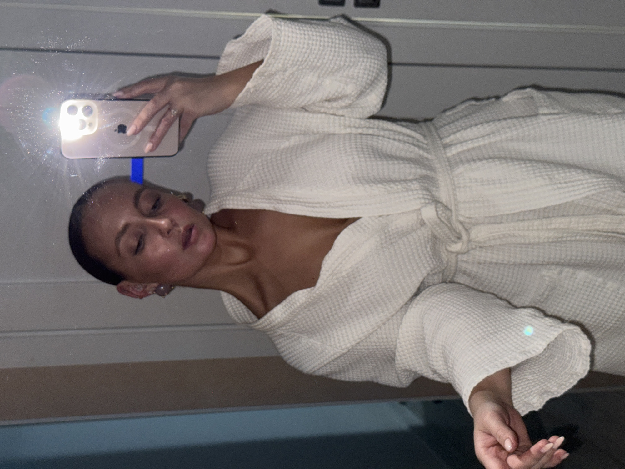 A woman in a white waffle-textured bathrobe is taking a selfie in a bathroom mirror. The phone flash is prominently visible. The location is likely a bathroom in Chaikovsky, Russia, but this cannot be definitively confirmed from the image.