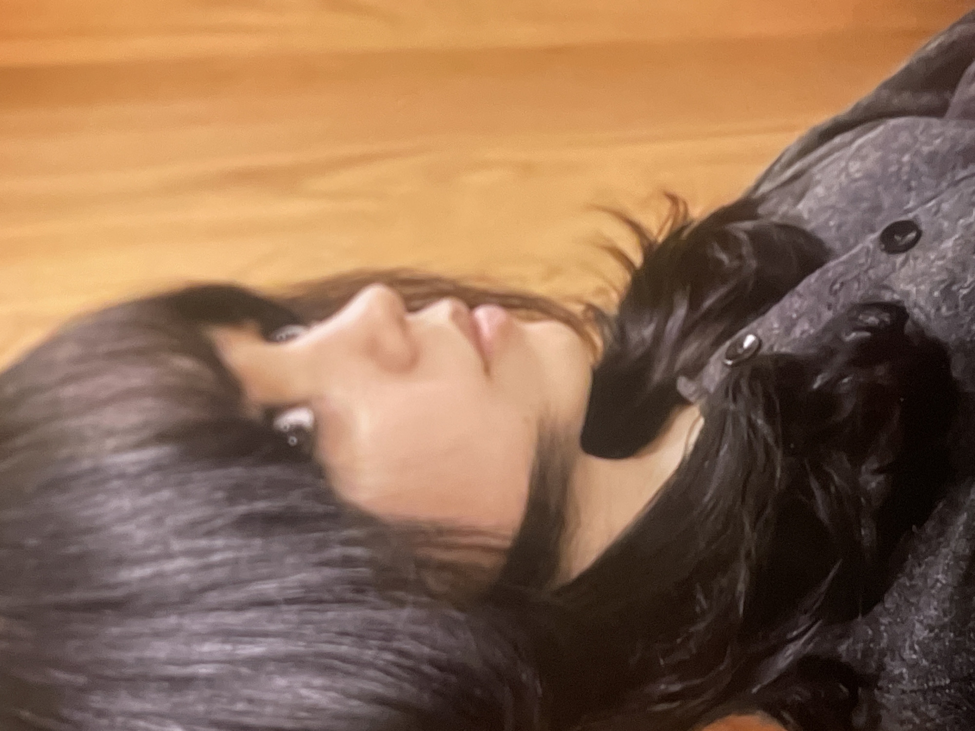 The image presents a close-up, angled view of a person, likely a young female, with long, dark hair, positioned diagonally across the frame. Her head rests on a light brown surface, possibly a wooden floor or wall, indicating an indoor setting.
The subject's face is seen in profile, revealing light skin, an open eye, a small nose, and slightly parted lips, conveying a calm or thoughtful expression. Her dark hair, which appears to be black or very dark brown, frames her face and spreads out behind her head.
She is wearing a dark, possibly grey or black, textured garment, which appears to be a jacket or coat, with visible dark buttons or embellishments on the shoulder or upper arm area. The focus is primarily on her face and hair, with the background being a soft, uniform light brown. There is no visible text in the image. No specific activity beyond lying down can be definitively determined. The time of day and weather are not discernible. While the location context is provided as Poltava, Ukraine, there are no visual cues within the image to support this.