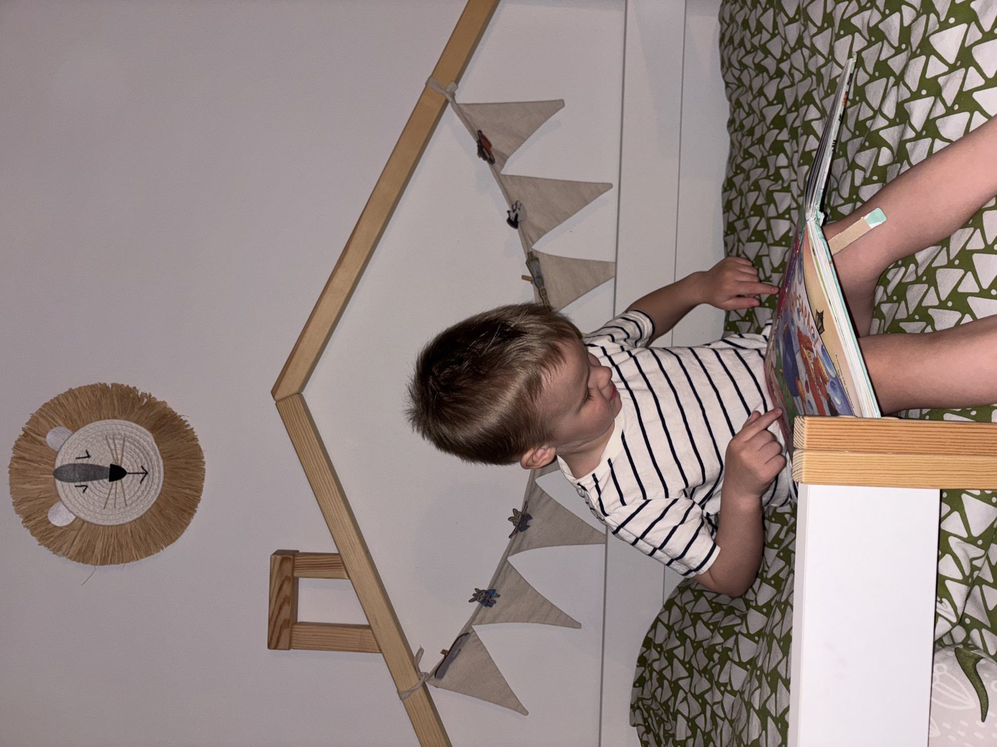 A young child with short blonde hair wearing a striped shirt sits in a bed with green patterned bedding, reading a book. The bed frame is white with light wood trim. Above the bed, a decorative wooden frame in the shape of a house is visible, adorned with small triangular flags. On the wall, a woven lion decoration hangs. The image was taken indoors. It is not possible to determine the location within Chaikovsky, Russia from the image alone.