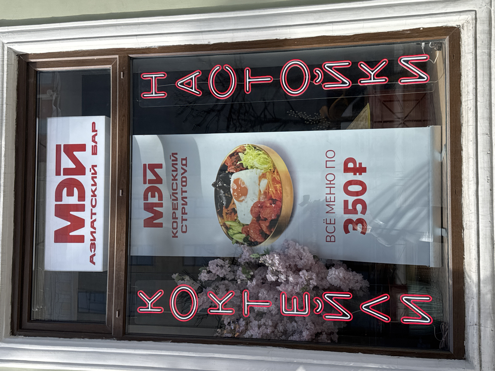 The image shows a storefront window for a Korean restaurant/bar called "Mey" in what appears to be Saint Petersburg, Russia. The sign is in Russian with some of the letters in the name spelled out backward. The window display includes signs in Russian advertising the restaurant's name ("МЭЙ"), its cuisine ("КОРЕЙСКИЙ СТРИТФУД" - Korean street food), and the fact that a full meal costs 350 rubles. There is an image of a Korean dish, likely bibimbap, and artificial flowers at the bottom of the window.