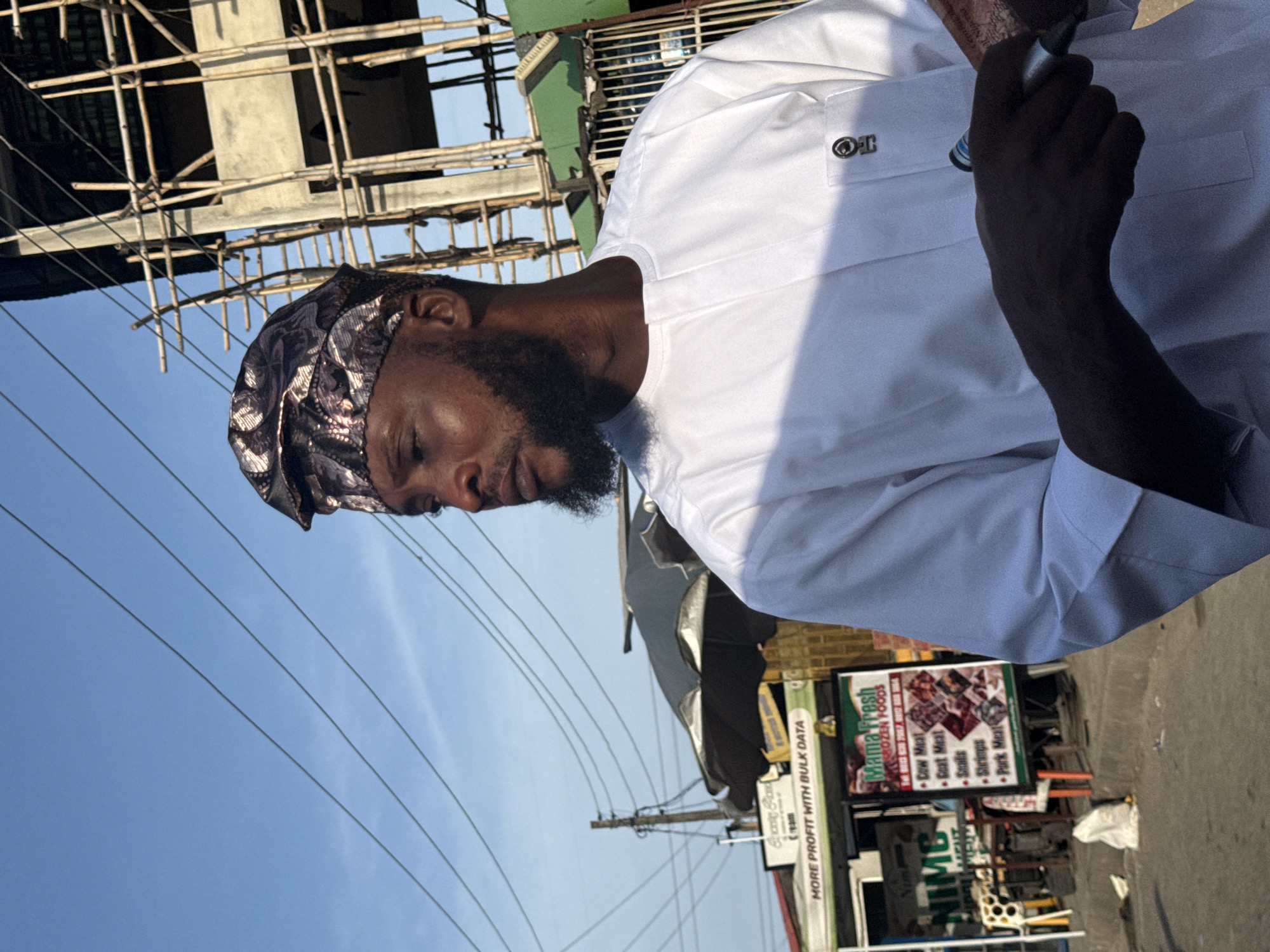 The image captures a street scene in Ado, Nigeria, featuring a man in the foreground against a backdrop of commercial activity and construction.
The primary subject is a dark-complexioned man, likely in his 20s or 30s, with a full, neatly trimmed dark beard. He is wearing a white, long-sleeved traditional tunic, possibly made of cotton, with a simple round neckline. On the left breast pocket of his tunic, a small circular emblem is pinned, featuring what appears to be a stylized "TO" or similar monogram. His head is covered by a patterned cap (fila), which is dark brown, black, and bronze/gold in color, with intricate abstract designs. He is positioned facing slightly right of the viewer, looking towards his left with a neutral expression. His right hand, partially obscured by shadow and holding a dark cylindrical object (possibly a marker or pen), is also holding several Nigerian currency notes, one of which clearly displays "Central Bank of Nigeria" text and appears to be a reddish-brown denomination (e.g., 200 Naira or 500 Naira).
In the mid-ground to the left, a series of makeshift market stalls or shop fronts are visible. A green sign with white text reads "GLOCLAW" and "MORE PROFIT WITH BULK DATA." Adjacent to it is a larger sign with a dark frame, white background, and red text, advertising "Mama Fresh FROZEN FOODS" and listing products such as "Cow Meat," "Goat Meat," "Snails," "Shrimps," and "Pork Meat," accompanied by images of meat cuts. A large, dark market umbrella is open behind these stalls. Utility poles and multiple overhead power lines traverse the clear, light blue sky on the left side of the frame.
To the right of the subject, in the background, a multi-story building is under construction, characterized by extensive scaffolding made of wooden poles. The building appears to be concrete, with some sections showing exposed concrete and others hinting at green paint. The sky above is clear and bright blue, indicating good daylight conditions. The ground beneath the market stalls appears to be a paved surface.