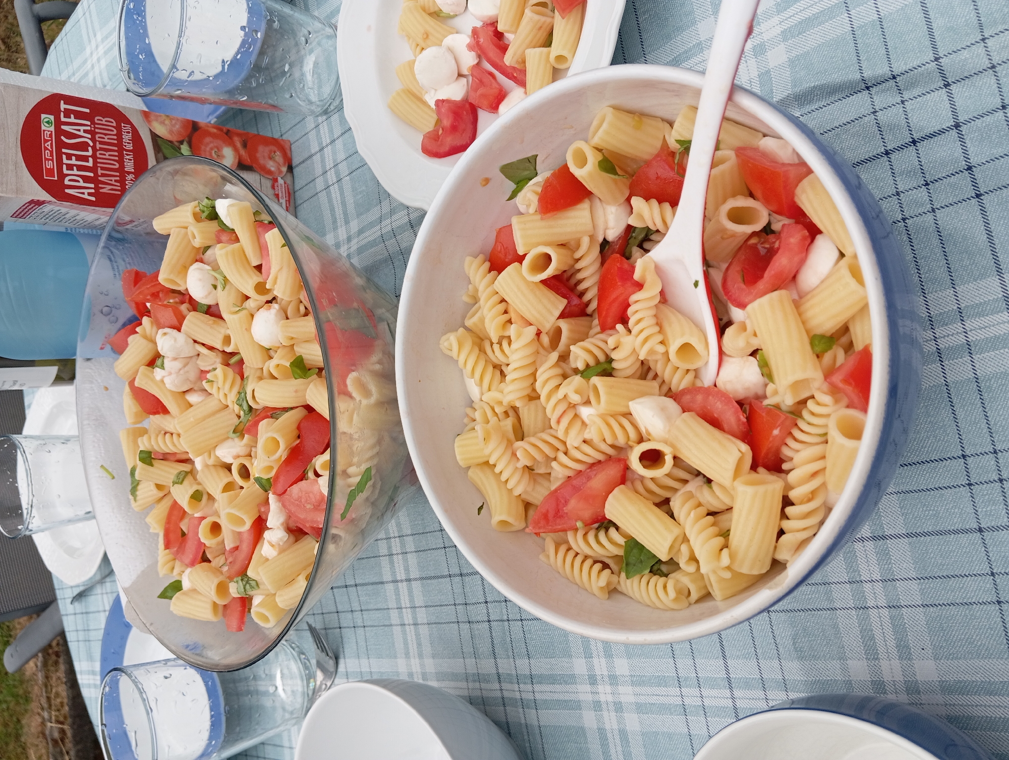 The image shows a picnic table with two bowls of pasta salad, one in a large glass bowl and the other in a white ceramic bowl. Both bowls contain pasta, tomatoes, and mozzarella cheese, and are topped with basil leaves. The table is covered with a blue and white checkered tablecloth, and there are glasses of water and a carton of apple juice on the table. The scene is set outdoors, with green grass and trees visible in the background. The lighting suggests it is daytime, likely late morning or early afternoon, with soft, diffused light. The overall atmosphere is relaxed and casual, indicative of an outdoor meal. The presence of water glasses and a juice carton suggests a meal or gathering is in progress. The image composition focuses on the food, highlighting the freshness of the ingredients. The table setting implies a domestic or recreational outdoor space.