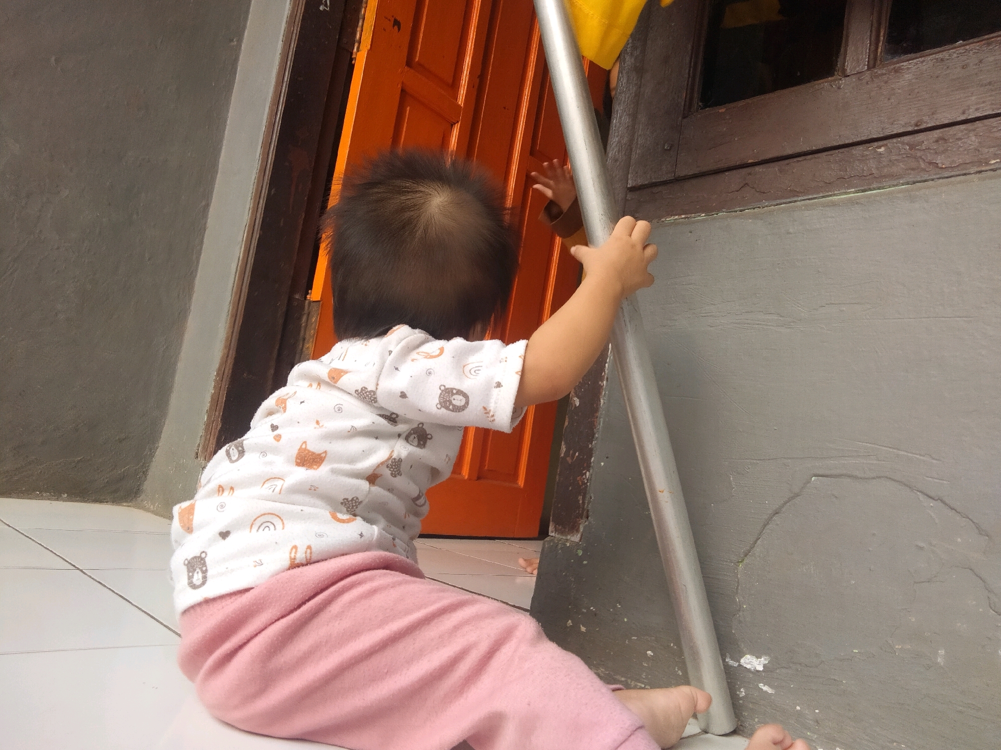 An overhead shot shows a baby from behind, wearing a white shirt with cartoon animal patterns and pink pants. The baby is reaching out to a metal pole. The background includes an orange door and a wooden window frame. The scene appears to be in a residential area, possibly a porch or entryway, in Suka Sari, Indonesia. The ground is tiled.