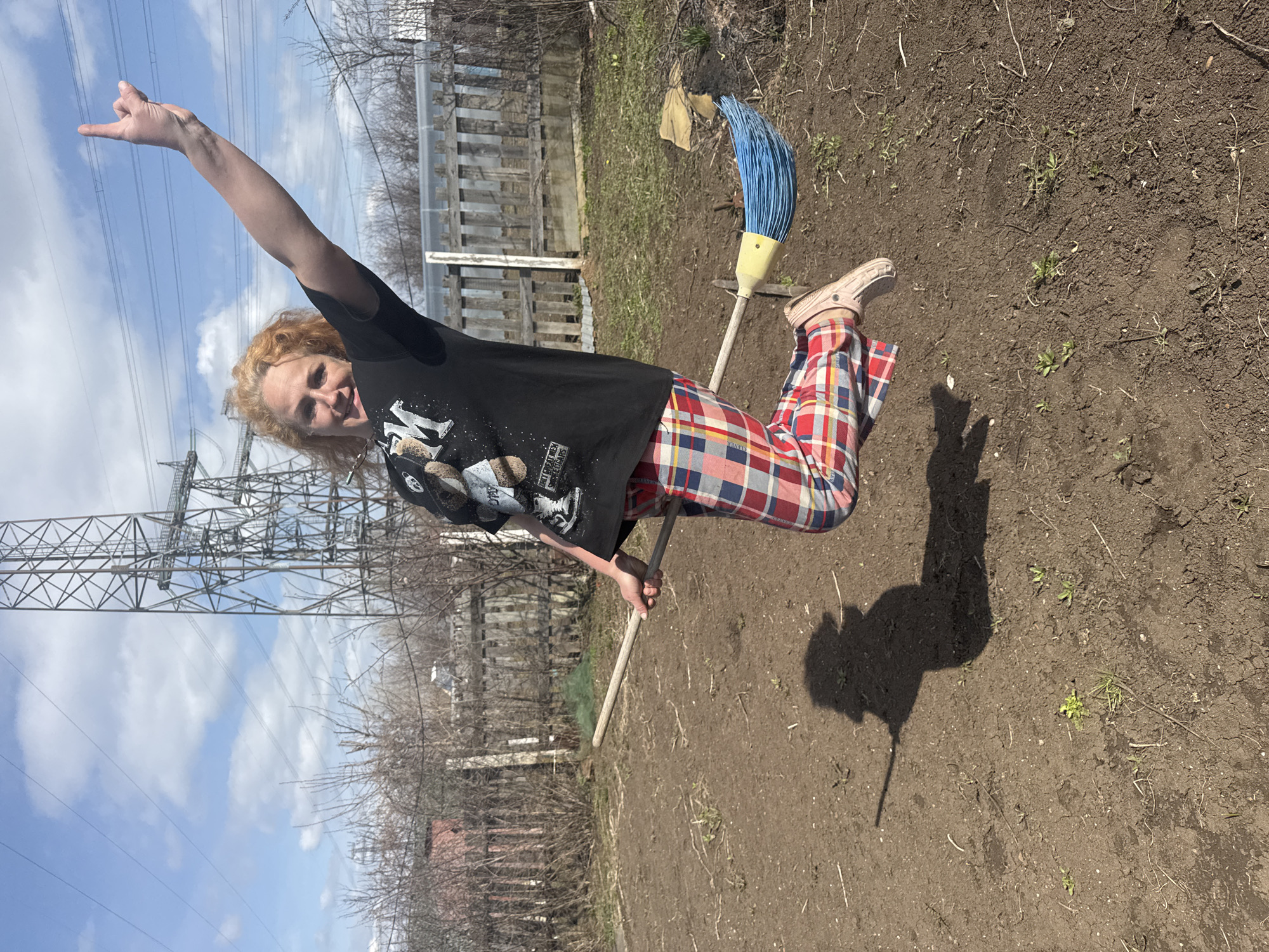 An outdoor, horizontally oriented full-shot captures a person posing playfully as if flying on a broomstick, set against a semi-rural backdrop in Chaikovsky, Russia. The individual, with reddish-blonde curly hair, is looking towards the camera with a wide, joyful smile, conveying a lighthearted and energetic mood.
They are wearing a black t-shirt with a graphic on the front that includes text like "THE GREAT AMERICAN" and other motifs. Their attire also features distinctive red, blue, white, and yellow plaid trousers, complemented by light-colored Crocs. The person's right arm is extended upwards, making a "rock and roll" hand gesture (corna), while their left hand grasps the wooden handle of a broom. The broom has bright blue bristles and a yellow plastic head, positioned between their legs to simulate flight.
A distinct shadow of the person and the broom is cast on the ground, which appears to be tilled or disturbed earth, with sparse green sprouts and dry vegetation. In the background, a rustic wooden fence is visible, along with bare trees and bushes, suggesting an early spring or late autumn season. A prominent feature on the left side of the frame is a large metal power transmission tower with multiple power lines stretching across the bright blue sky, which is dotted with white clouds. The strong sunlight indicates it is daytime.