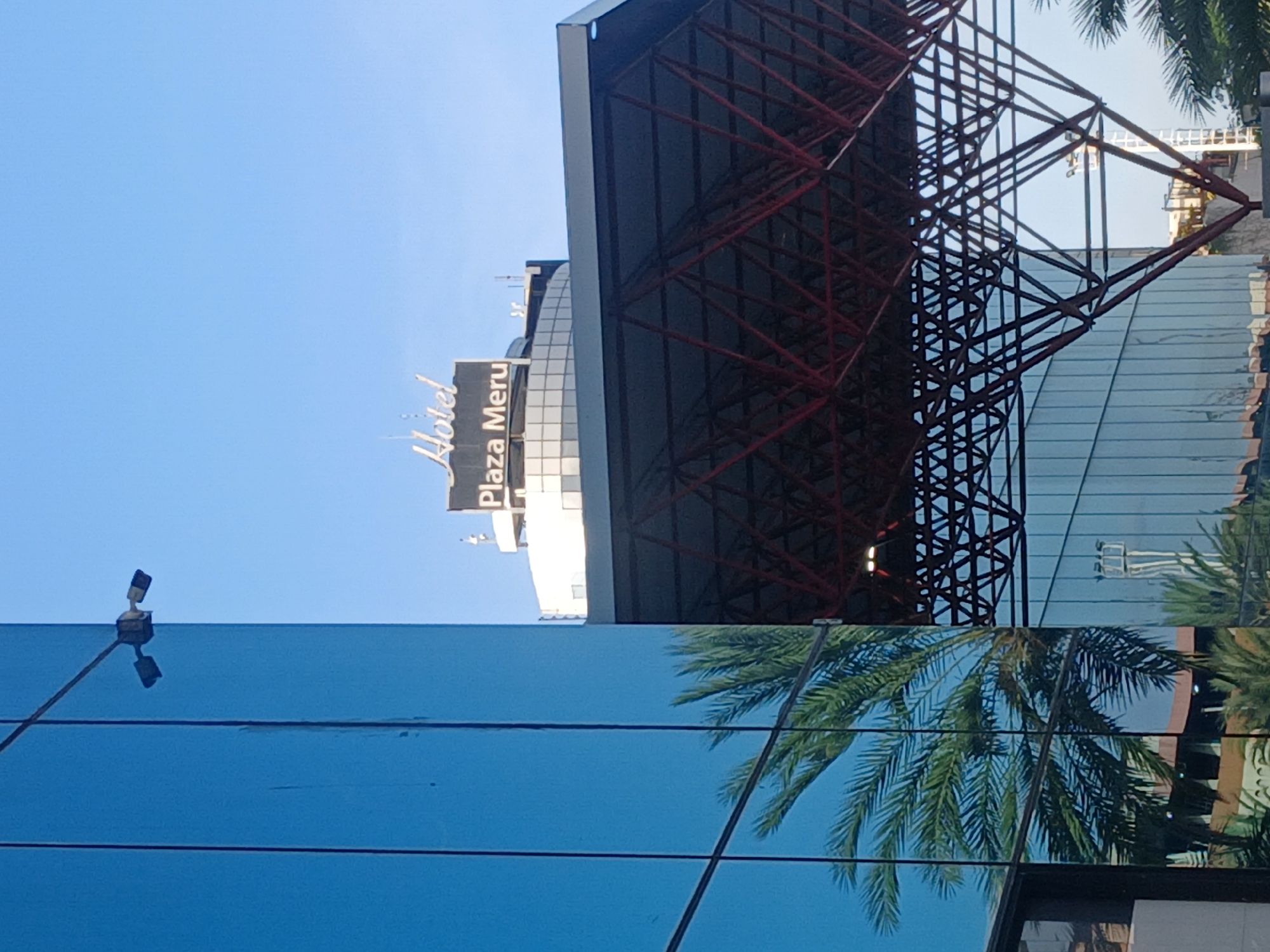 The image features the Plaza Meru Hotel in Ciudad Guayana, Venezuela. The hotel's signage is visible against a bright blue sky. A red and black metallic structure is prominent in the composition. The scene is reflected in a mirrored surface. A palm tree frond appears to be partially reflected. A street light or security camera is also visible on the left.
