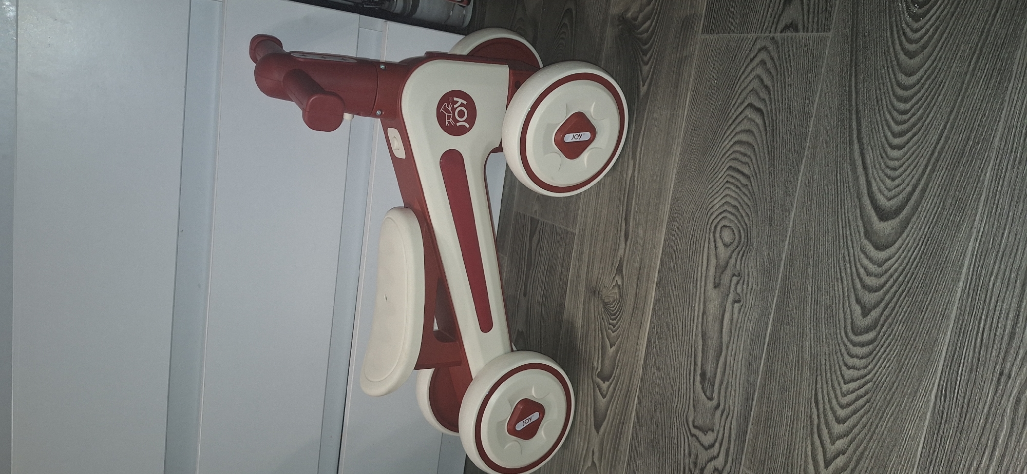 A red and white ride-on toy bicycle is placed on a wooden floor in front of a white cabinet. The toy has four wheels, a seat, and handlebars. The word "JOY" is visible on the side of the toy. The background appears to be indoors, likely a home environment. There are no people present in the image. The location of this scene in Bila Tserkva, Ukraine is not determinable from the image alone.