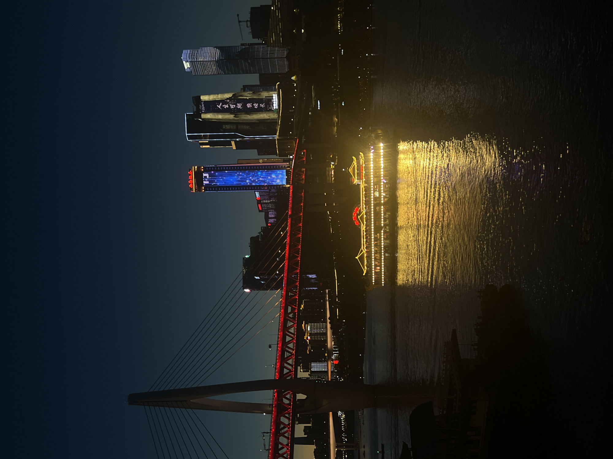 A long shot captures the cityscape of Yuzhong District, China, at dusk, illuminated by city lights and a sunset that has left a deep indigo sky. The foreground is dominated by a wide river, its surface reflecting the warm glow of the city. On the water, a traditional Chinese boat with a red roof and glowing yellow lights cruises, its reflection shimmering on the water.
In the mid-ground, a modern suspension bridge, with a single tower on the left, is dramatically lit with red lights that outline its structure and deck. The bridge spans the river, connecting different parts of the city. Behind the bridge, a cluster of skyscrapers rises, their facades adorned with colorful lights. The tallest building on the right displays a light show in blue, while the central one features golden Chinese characters.
The background consists of a dark, hilly terrain dotted with smaller buildings and lights, suggesting a densely populated urban environment. The overall scene depicts a vibrant, nocturnal urban landscape where modern architecture and traditional elements coexist.