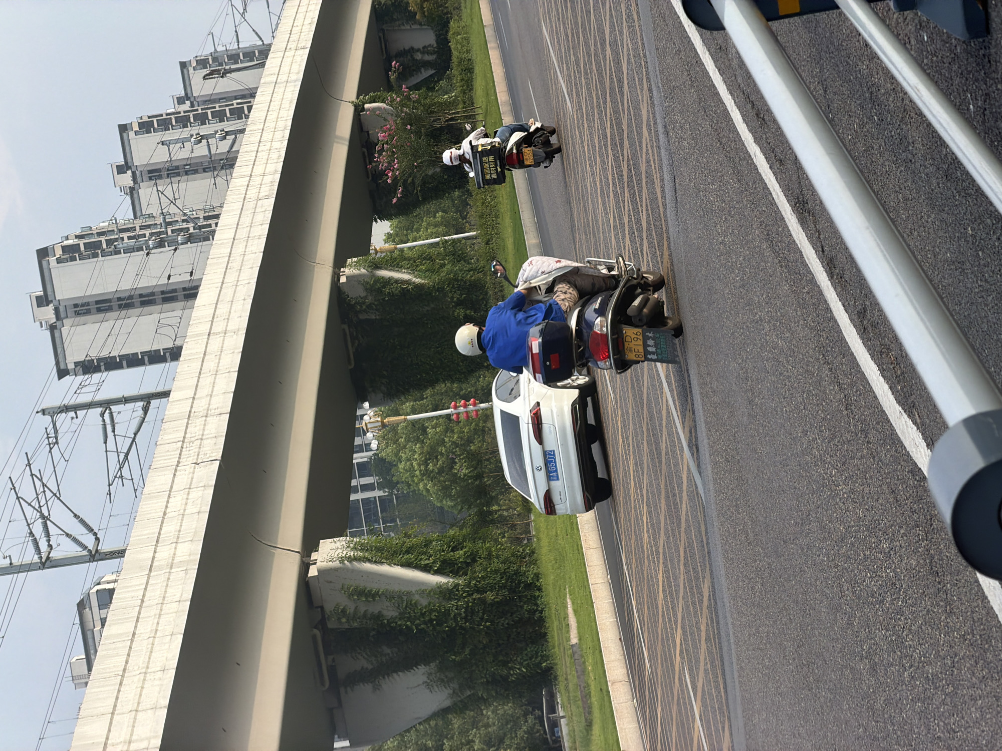 In Jiangjin, China, a concrete overpass stretches across a road with overhead power lines. Two individuals on scooters navigate the asphalt, one in a blue shirt and helmet, the other in white with a white helmet. A white Volkswagen sedan, bearing the license plate "A 65J72," is parked on the road. Lush green vegetation climbs the supports of the overpass, and modern buildings rise in the background. The scene is captured during daylight, under an overcast sky.