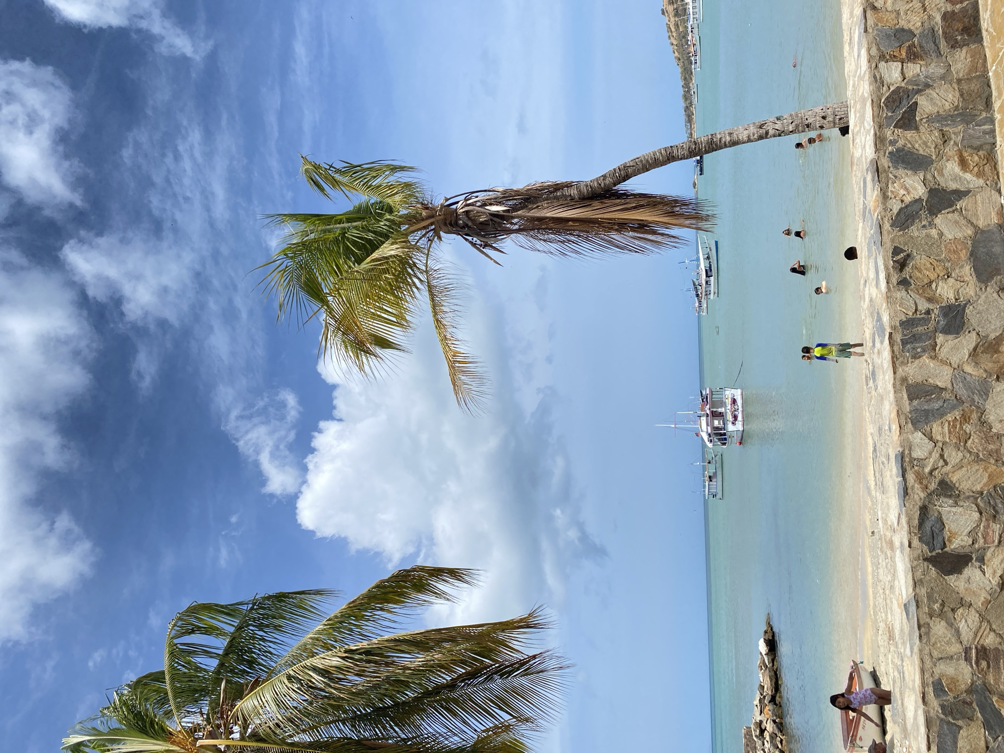 The image shows a beach scene in Juan Griego, Venezuela. Two palm trees are visible, with one bending towards the beach. Several people are in the water swimming, and boats are anchored offshore. A stone wall runs along the edge of the beach. A young girl stands with a small boat on the sand. The sky is blue with white clouds.