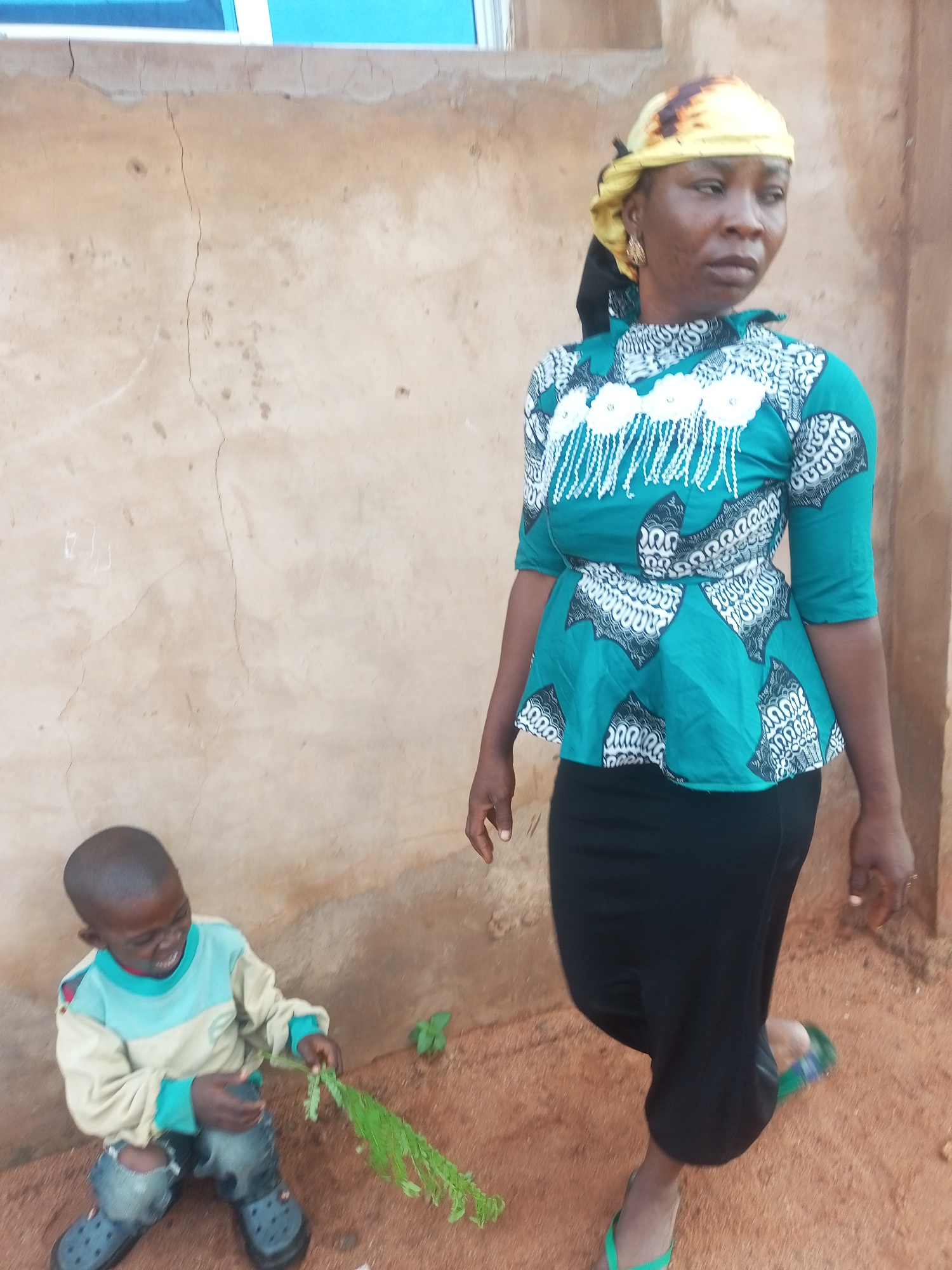 In Tempure, Nigeria, a woman and a child are captured in a full-body photograph. The woman, standing on the right, wears a vibrant teal peplum top adorned with a white, abstract pattern and fringes, paired with a black skirt and green flip-flops. Her head is covered with a yellow and brown patterned headscarf, and she has a gold earring in her left ear. She looks to her right, her expression unreadable. A young boy, crouched on the left, is dressed in a light blue and beige long-sleeved shirt with ripped denim jeans and dark grey crocs. He is holding a bunch of green leaves and appears to be smiling or laughing. The background consists of a textured, light brown wall with visible cracks and a hint of a blue window frame at the top. The ground is covered in reddish-brown soil.