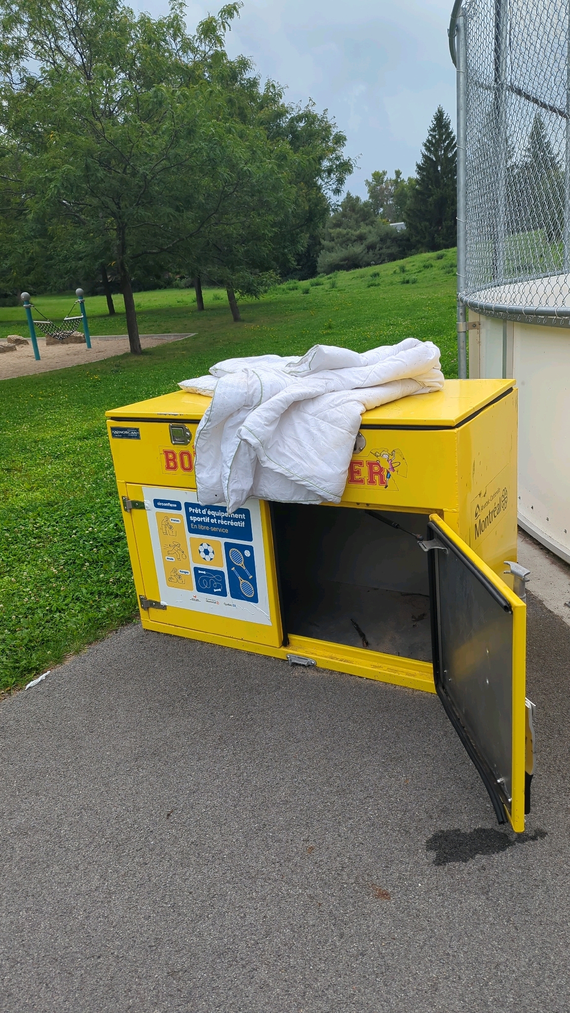 A white quilted blanket is draped over the top of a bright yellow, metal donation bin labeled "BOXER" in large red letters. The bin has a hinged door on the front that is open, revealing a dark interior. A colorful decal on the left side of the bin shows icons of sports equipment and text that reads "Prêt d'équipement sportif et récréatif En libre-service". The bin is situated on a paved surface next to a grassy area with trees. In the background, a chain-link fence encloses what appears to be a sports field or court. The overall scene suggests an outdoor park or recreational area in Montreal, Canada, with the bin likely intended for donations of sports and recreational equipment.