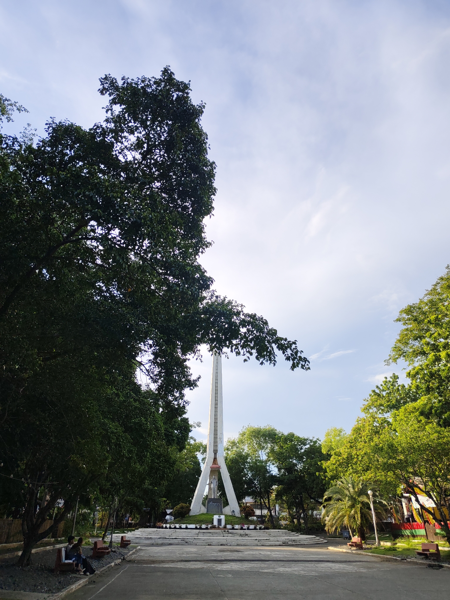 The image depicts a monument in Davao City, Philippines, situated in a park-like setting. The monument, a tall, slender obelisk with a triangular base, stands prominently in the background. At its base, a statue of a figure in military uniform is visible. Surrounding the monument are mature trees with lush green foliage, some of which extend into the foreground, partially obscuring the sky.
In the foreground, a paved area leads to the monument. Two people are seated on a bench on the left side of the image, facing away from the monument. The bench is made of wood and appears to be placed on a bed of gravel. Other benches are visible in the middle ground, some empty and some with people seated. A lamppost stands to the right of the monument, illuminated by the natural light. The sky above is a pale blue with scattered white clouds, suggesting it is daytime. The overall scene conveys a sense of peace and serenity in a public space.