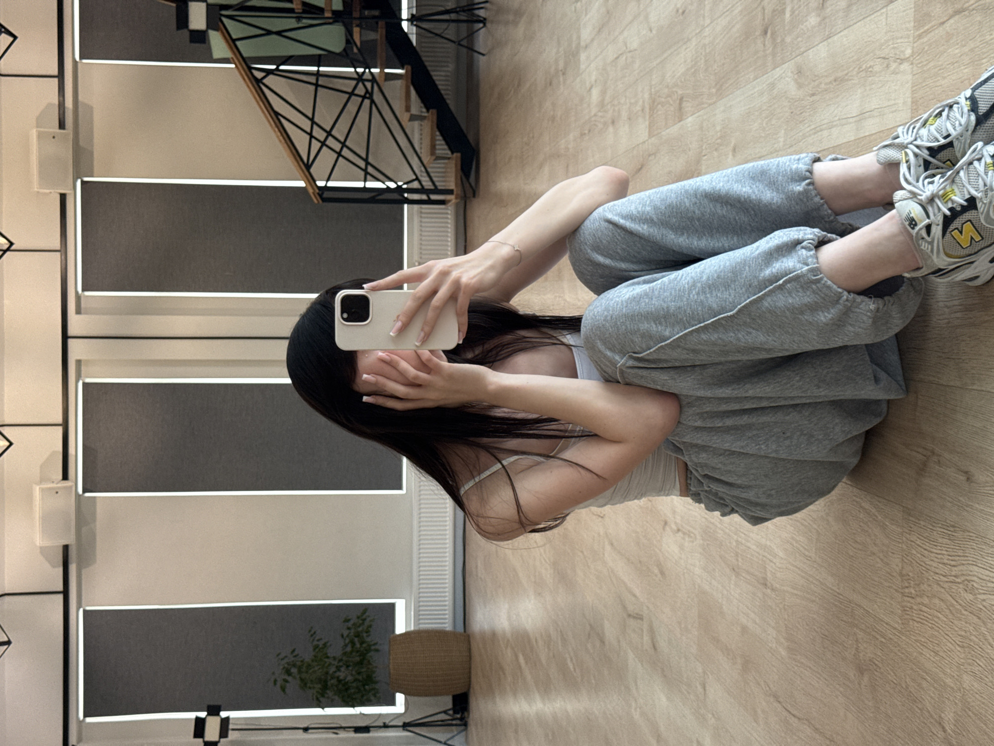 The image shows a young woman sitting on a wooden floor, taking a selfie with her phone. She is wearing a white tank top and grey sweatpants. Her long, dark hair covers part of her face, and she has her hands raised to her face, obscuring her eyes. She is wearing white sneakers with grey and yellow accents.
The setting appears to be an indoor space with a modern aesthetic. The floor is light-colored wood. In the background, there are large windows with blinds, and one section of the wall has a metal geometric design. A black staircase with a railing is visible in the upper left corner, reflecting in a mirrored surface. A woven basket and a plant are also visible in the background. The lighting suggests it is daytime, possibly late afternoon or early evening, based on the indoor lighting and the soft illumination. The overall mood is casual and relaxed. There is no visible text in the image. The location context provided is Zakharovo, Russia.