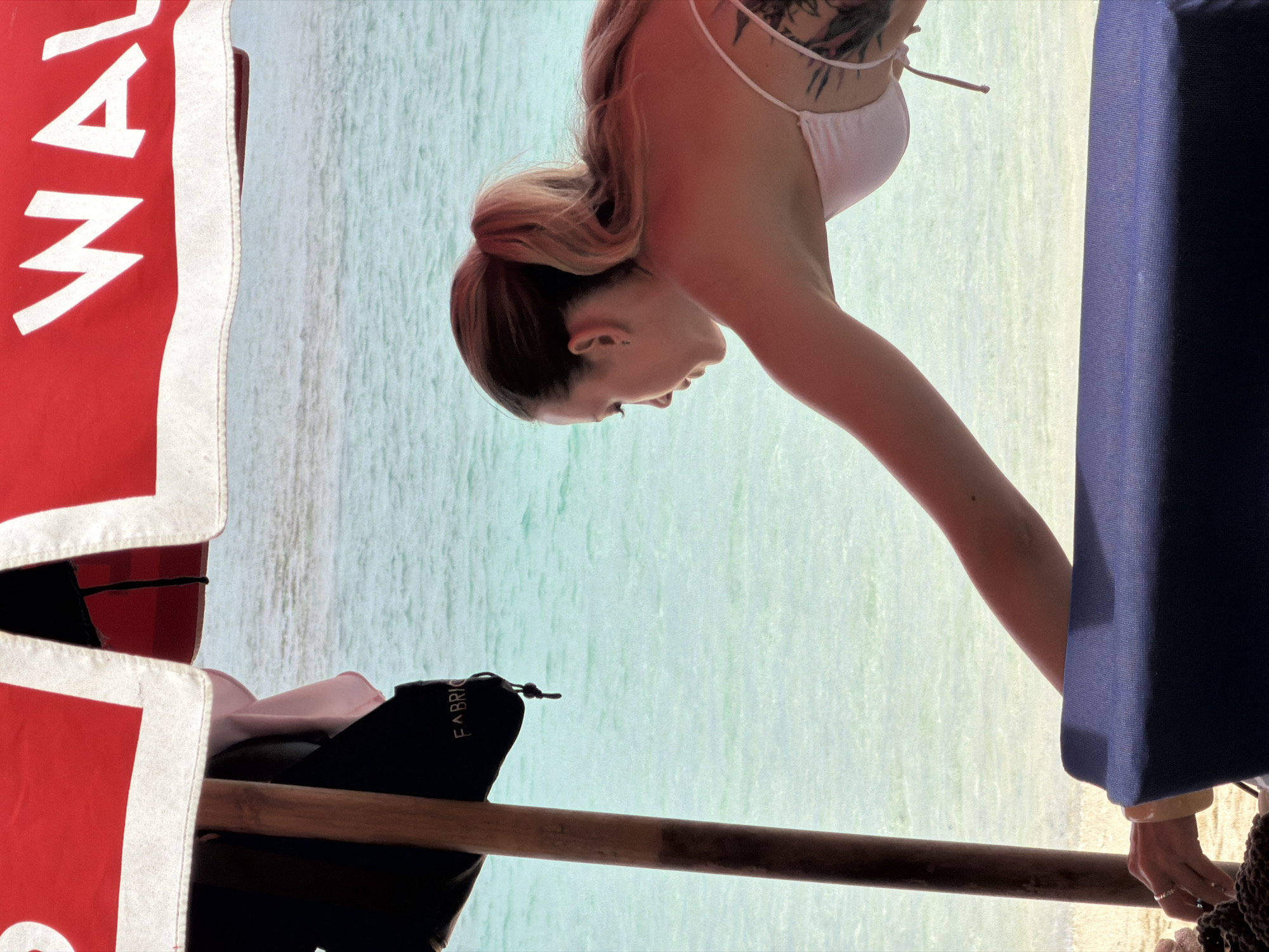 A woman in a white bikini is leaning forward, reaching towards the viewer's left, with her right arm extended. She has blonde hair pulled back in a ponytail and a tattoo on her back and shoulder. She is positioned on a blue cushioned surface, possibly a beach chair, with a wooden pole to her left supporting a red and white striped umbrella or awning. The background consists of clear, shallow turquoise water with a sandy bottom visible. Text on the umbrella reads "WAL". A black pouch is hanging from the wooden pole.