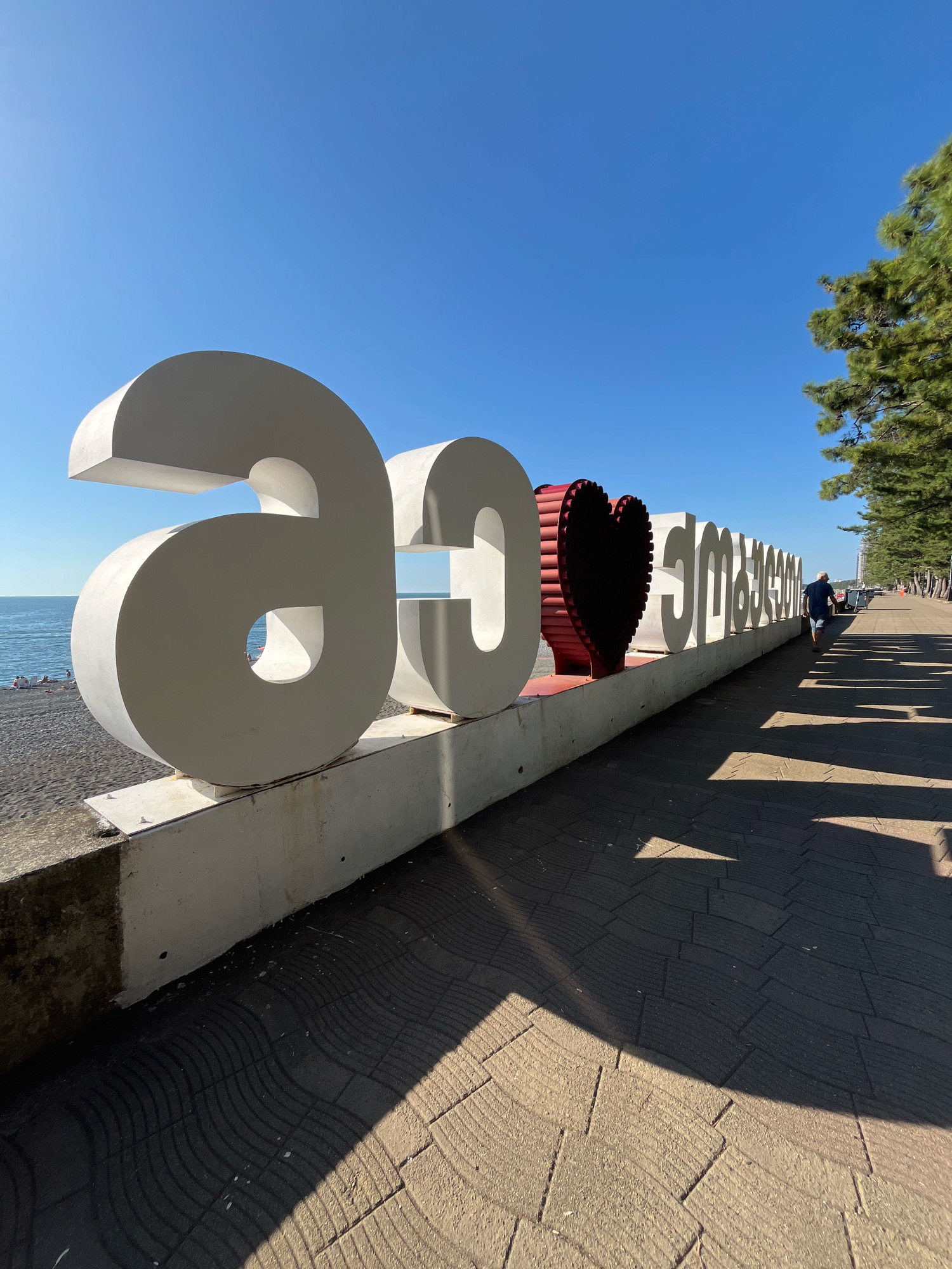 A wide-angle, eye-level, outdoor shot captures a large, white sculpture spelling out "I love Kobuleti" in Georgian script against a clear blue sky. The sculpture, situated on a concrete base along a paved promenade, features large, white letters with the exception of the heart-shaped middle character, which is red with a corrugated texture. To the left of the sculpture is a dark pebble beach and the calm blue sea. To the right, a tree-lined promenade stretches into the distance, casting long shadows from the sculpture. A lone person in a dark blue shirt and shorts is walking away from the camera on the promenade, and a few people are visible on the beach in the background. The lighting suggests it is daytime, with the sun casting sharp shadows.