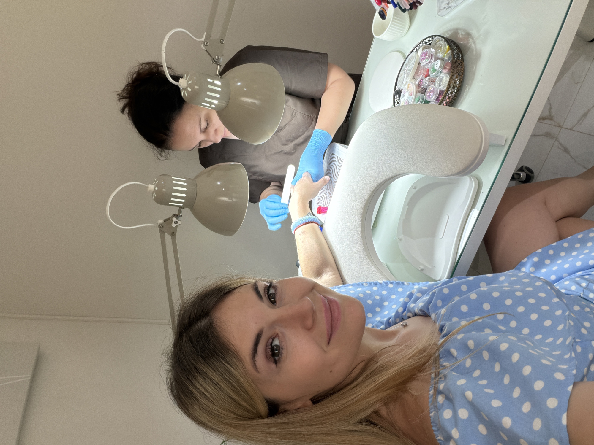 The image depicts two women in what appears to be a nail salon. One woman, with dark hair pulled back, is wearing a brown top and blue gloves, is working on the nails of another woman wearing a blue and white polka dot top. The second woman, facing the camera, has blonde hair and is smiling. Two lamps are positioned to illuminate the work area, which includes a white desk, a nail treatment station with an armrest, and small decorative containers. The location is potentially Budva, Montenegro.