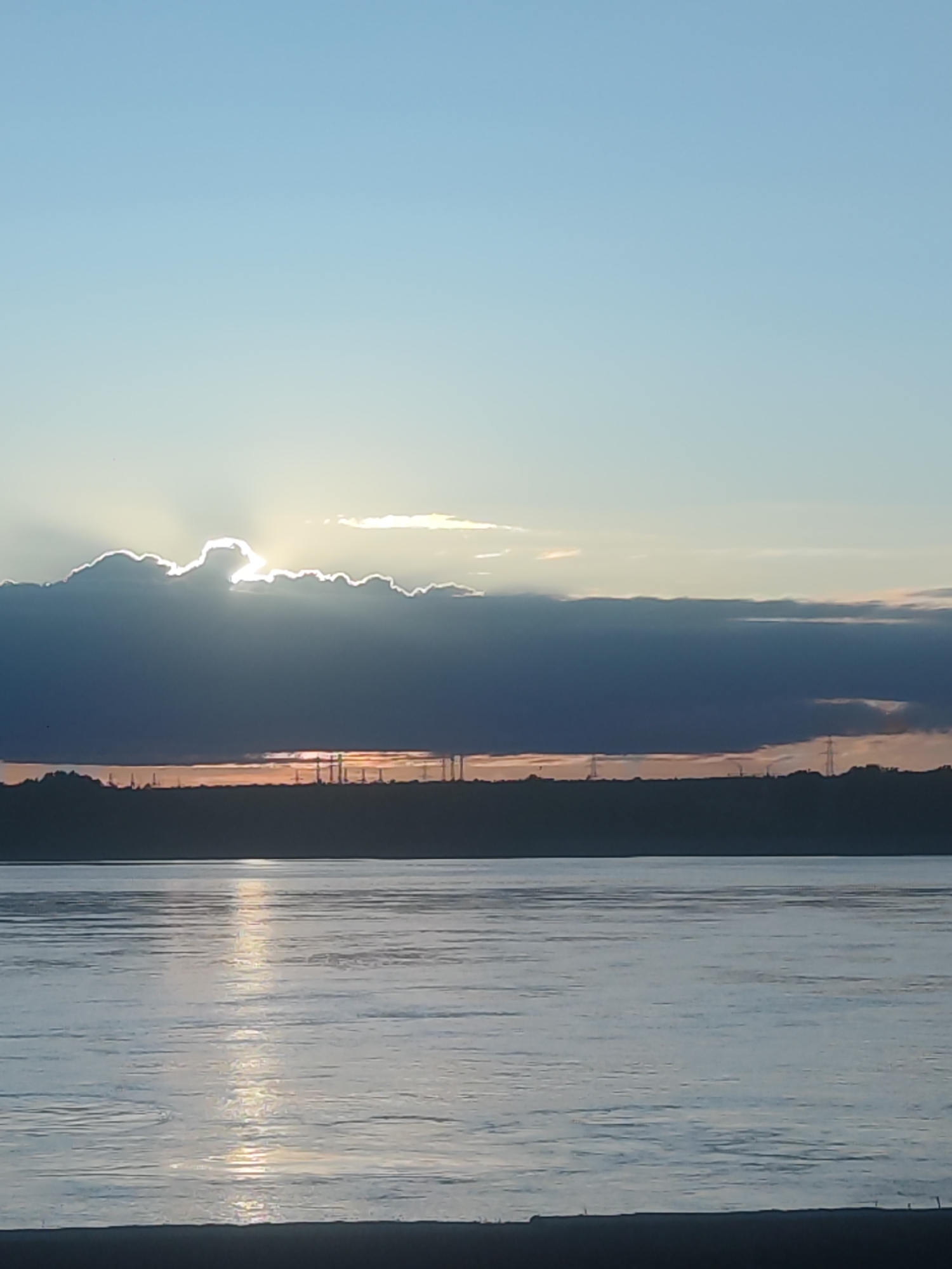The image presents a serene waterscape at either sunrise or sunset, dominated by a large body of water in the foreground and a dramatic sky. The upper third of the frame shows a clear, light blue sky, transitioning to a pale yellow-orange near the horizon. A thick, dark band of clouds spans the middle of the sky, with the sun partially hidden behind it. Bright rays of sunlight burst through a gap in these clouds, creating a brilliant halo effect and radiating upwards.
Below the main cloud formation, a dark, silhouetted strip of land marks the distant shore. Along this horizon, several faint, vertical structures, possibly power line towers or industrial elements, are visible against the soft light. The lower half of the image is filled by a wide body of water, likely a river or lake, exhibiting subtle ripples. A prominent path of sunlight shimmers on the water's surface, reflecting the bright light from the sky and extending towards the viewer.
The scene conveys a peaceful atmosphere, emphasizing the interplay of light and shadow during a transitional time of day. There are no people or explicit activities depicted. No text is visible. The location is generic, characterized by water, land, and distant structures.