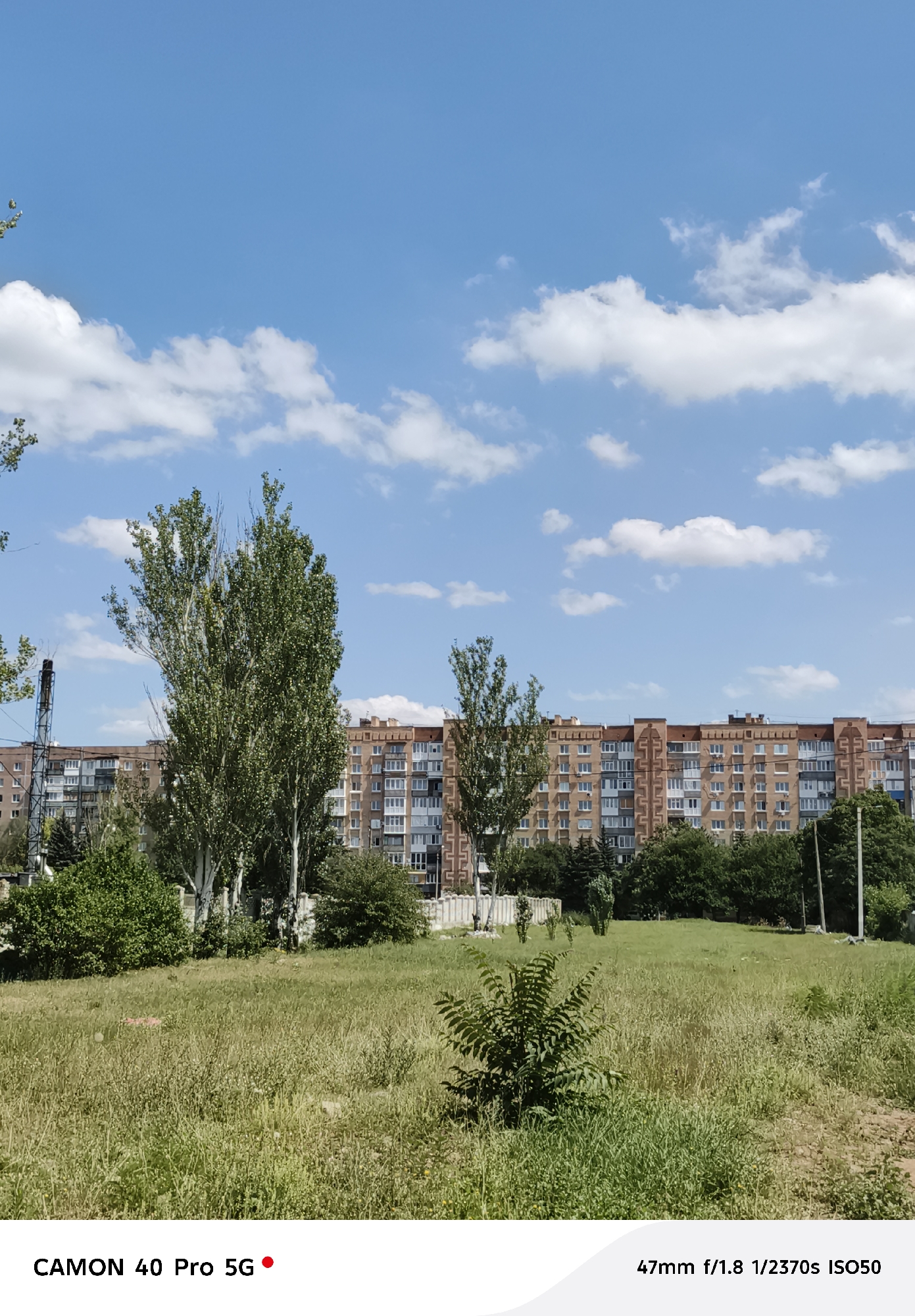 The image depicts a wide-angle outdoor scene under a bright blue sky with scattered white clouds. The foreground is a grassy field with tall, dry grass and a few small bushes. In the middle ground, several tall, leafy trees stand prominently, with a tall industrial structure visible to the left of the trees. Behind the trees and foliage, a long, multi-story apartment building stretches across the frame. The building has a brick facade and numerous windows, suggesting a residential area.
The scene appears to be set during the daytime, likely mid-morning or afternoon, given the strong sunlight casting shadows and the clear sky. The weather seems pleasant and sunny. While no people are visible, the apartment building indicates a populated area, likely a city or town. The presence of the apartment building and the overall urban-residential landscape could suggest a location within a city like Kramatorsk, Ukraine.
At the bottom of the image, there is visible text: "CAMON 40 Pro 5G" on the left and "47mm f/1.8 1/2370s ISO50" on the right. This text indicates the camera and some of its settings used to capture the photograph. There are no specific events or activities depicted, but the image captures a moment of a typical day in an urban environment.
