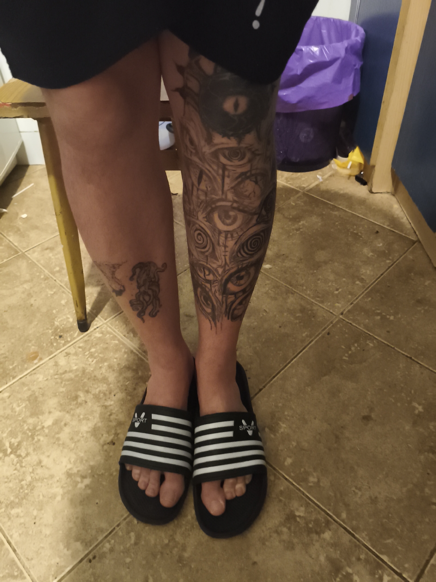 A person stands indoors on a tiled floor, their legs visible from mid-thigh down. They are wearing black shorts with a white exclamation point on the side, and black and white striped sandals labeled "SPORT". The right leg has a large tattoo featuring multiple eyes. The left leg has a smaller tattoo of an animal, possibly a tiger or panther. Behind the person are a yellow chair, a purple garbage can, and a blue wall. The image does not provide enough information to ascertain the precise location within Nowy Tomyśl, Poland.