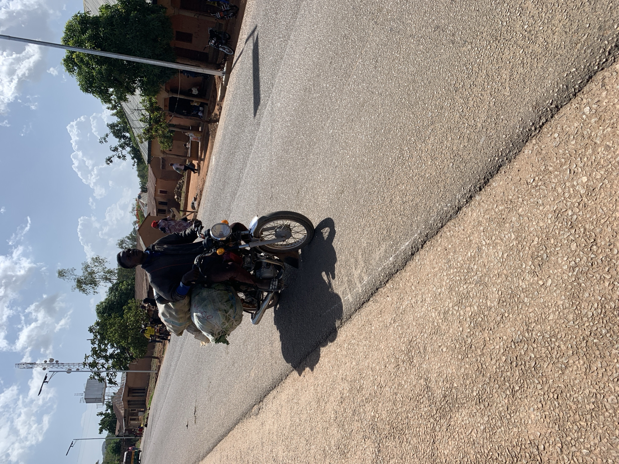 A man rides a motorcycle on a paved road in Vom, Nigeria. The motorcycle is laden with large sacks of goods. The road is lined with buildings and trees. In the background, there's a communications tower and other people are standing near some of the buildings. The sky is cloudy.