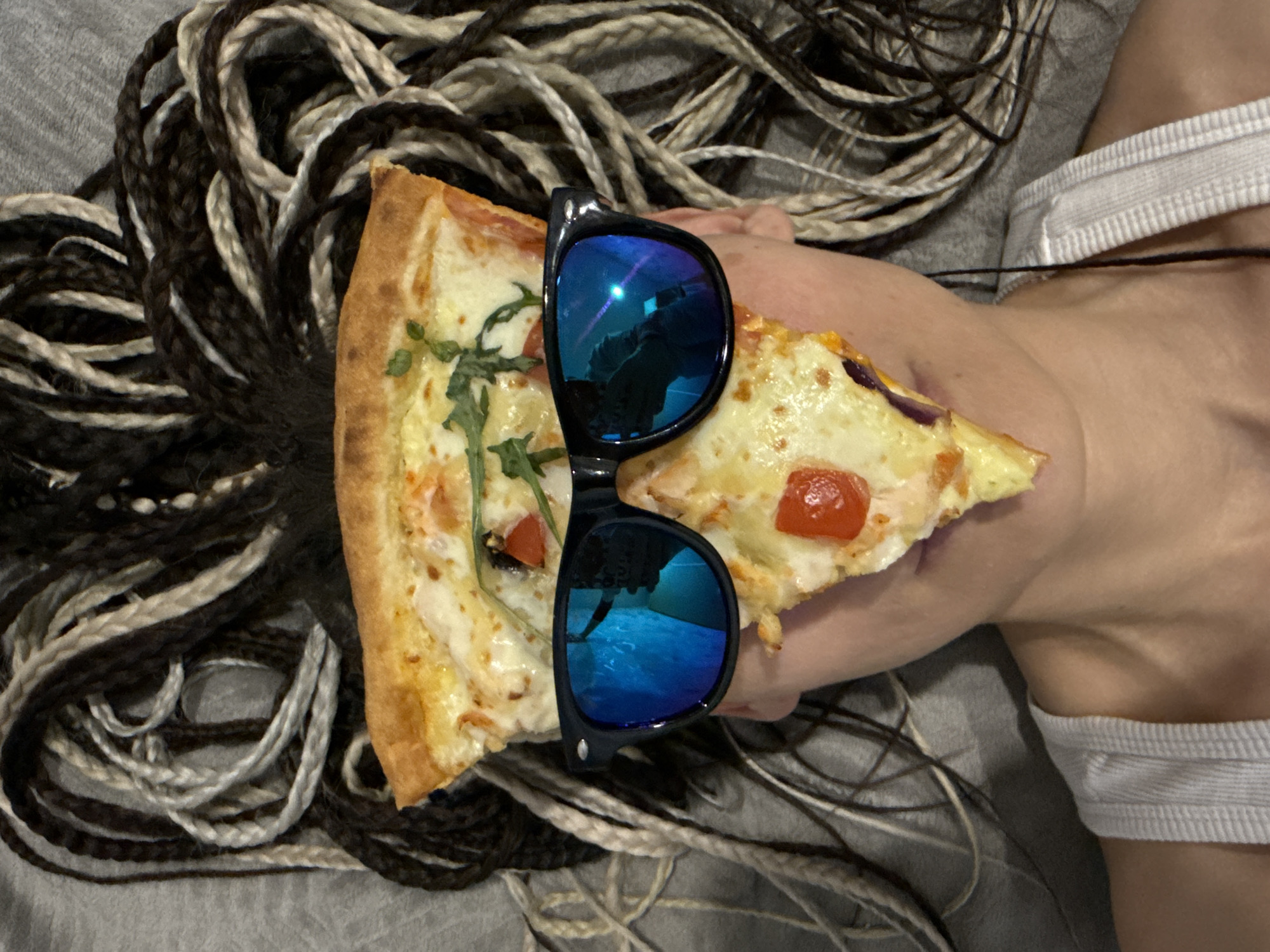 A close-up, top-down photograph captures a person holding a slice of pizza, presumably indoors in Rusanovo, Russia. The person's face is not visible, but their right arm, shoulder, and upper chest, wearing a white ribbed garment, are seen on the right side of the frame. Their distinctive hair, styled in numerous long, thick braids of alternating dark brown/black and light (possibly gray or bleached) colors, is spread out across a soft, gray fabric surface, suggesting a bed or couch.
The main focus is a large slice of pizza, held by the person's hand. The pizza features a golden-brown crust, melted cheese, bright red cherry tomatoes, and green herbs, possibly arugula. Resting on top of the pizza slice is a pair of black sunglasses with highly reflective, vivid blue lenses. The reflections in the sunglasses show distorted light sources or the ceiling, but no identifiable objects or text. The overall atmosphere is casual and relaxed. No text is visible in the image.