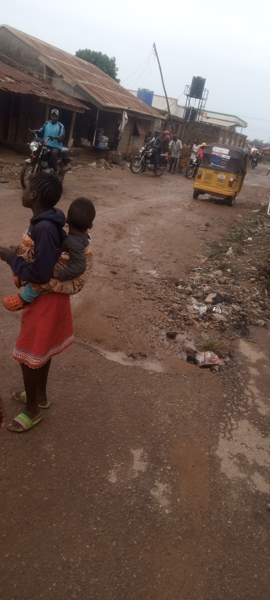 A young child carries a younger child on her back in a dirt road in Unknown, Unknown. The carrier wears a dark blue long-sleeve shirt, a red skirt with white and tan trim, and striped sandals. The carried child wears a gray shirt and orange shoes. They are secured by a patterned wrap. In the background, motorcycles and a yellow auto rickshaw travel on the dirt road, and people stand near buildings constructed with corrugated metal roofs. Trash litters the side of the road.