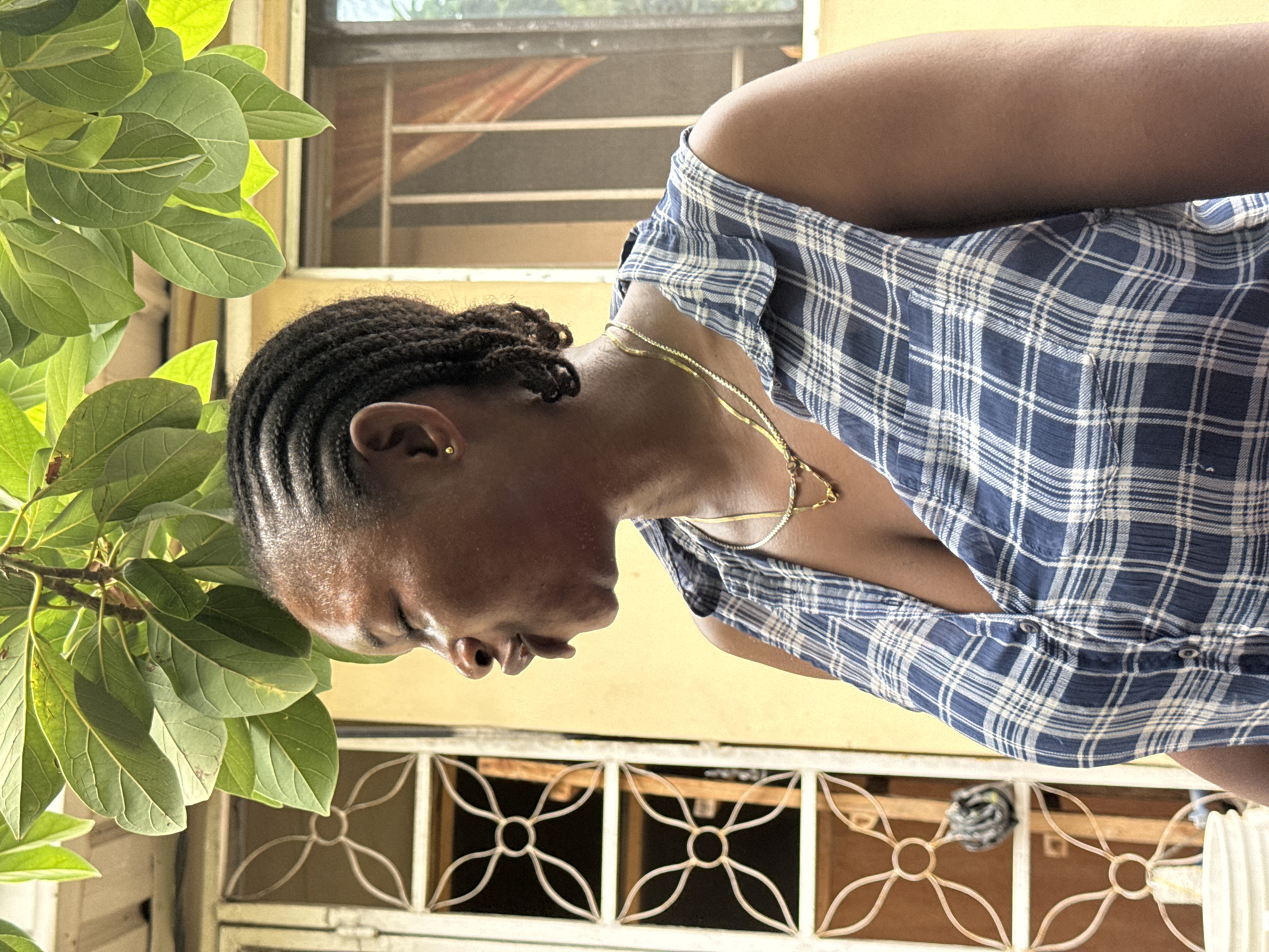A medium shot depicts a Black woman with dark braided hair, wearing a blue and white plaid shirt. She is facing downward and slightly to the left. The woman is wearing a delicate gold necklace. A plant with green leaves is to her left. Behind her is a window with a patterned metal security grate. The setting appears to be outdoors, possibly a yard. It is not possible to determine the precise location in Ado, Nigeria from this image.