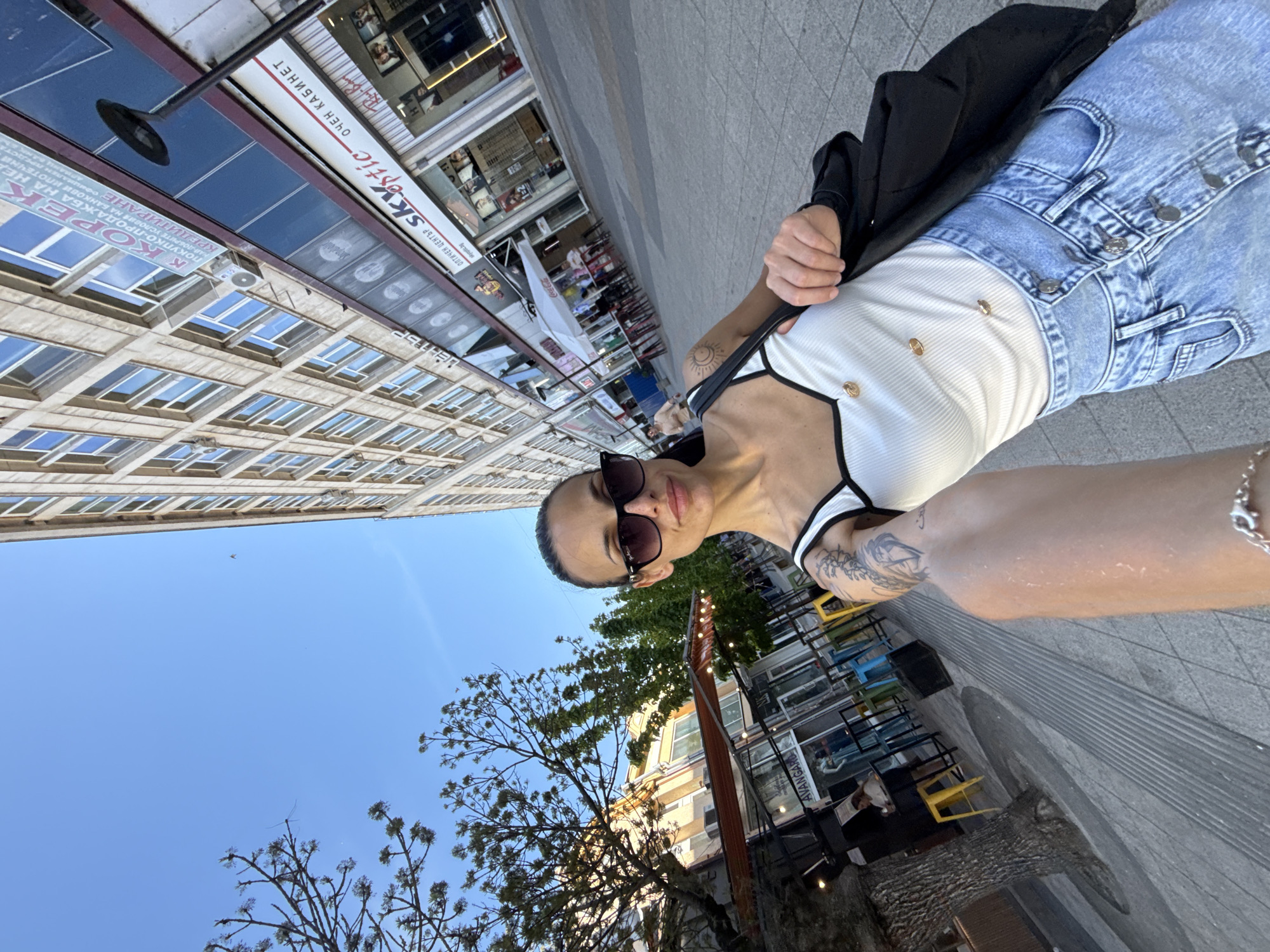 An individual is captured in a street-level perspective, appearing to take a selfie in an urban setting in Pleven, Bulgaria. The person, positioned in the foreground and slightly off-center, is wearing dark sunglasses, a white ribbed tank top with black trim and gold buttons, and light blue ripped jeans. A black jacket is casually draped over one shoulder. Notable tattoos are visible on their left arm, including a sun motif on the inner bicep and an intricate design on the forearm. Their expression is neutral to slightly smiling, partially obscured by the sunglasses.
The background reveals a bustling city street lined with multi-story buildings. The architecture features numerous windows, suggesting commercial or residential units above ground-floor businesses. Along the street, several shops are visible with awnings and signage. Visible text on the shop signs includes "skyeptic," "ОПТИЧЕН ЦЕНТЪР" (Optical Center), and "ОЧЕН КАБИНЕТ" (Eye Cabinet), indicating an optometrist or optical shop. Another sign promotes "ПОКУПКО-ПРОДАЖБА КБА НА НЕД" (Purchase-Sale KBA on Real Estate) and mortgage services, suggesting a financial institution. The brand "Coca-Cola" is also discernible.
Outdoor seating with colorful chairs (yellow, blue) and tables is arranged outside what appears to be a cafe or restaurant. Bare-branched trees with some green foliage are interspersed along the street, set against a clear, bright blue sky with a faint bird or distant object. The lighting indicates a sunny daytime, possibly late afternoon, given the shadows present. The overall scene depicts a lively commercial area.