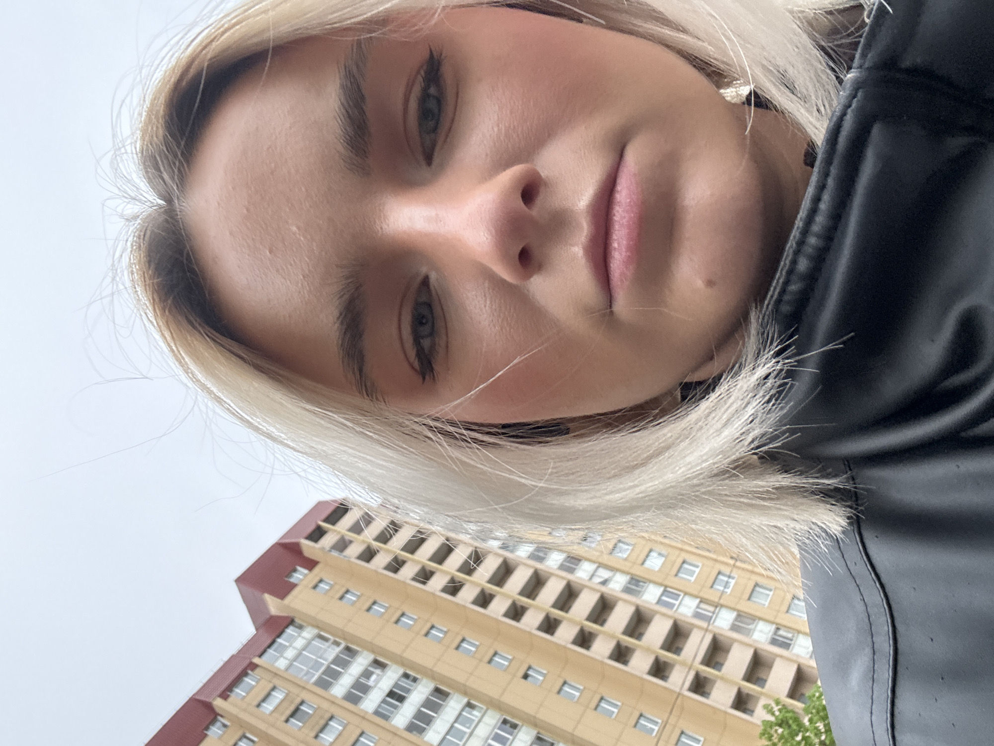 A photograph taken from a low angle features a young woman with blonde hair, wearing a black jacket. She looks directly at the camera with a neutral expression. Her makeup is visible, including eyeliner and blush. In the background, a multi-story building is partially visible. Based on the architecture, this photograph could have been taken in Saint Petersburg, Russia. The sky appears overcast.