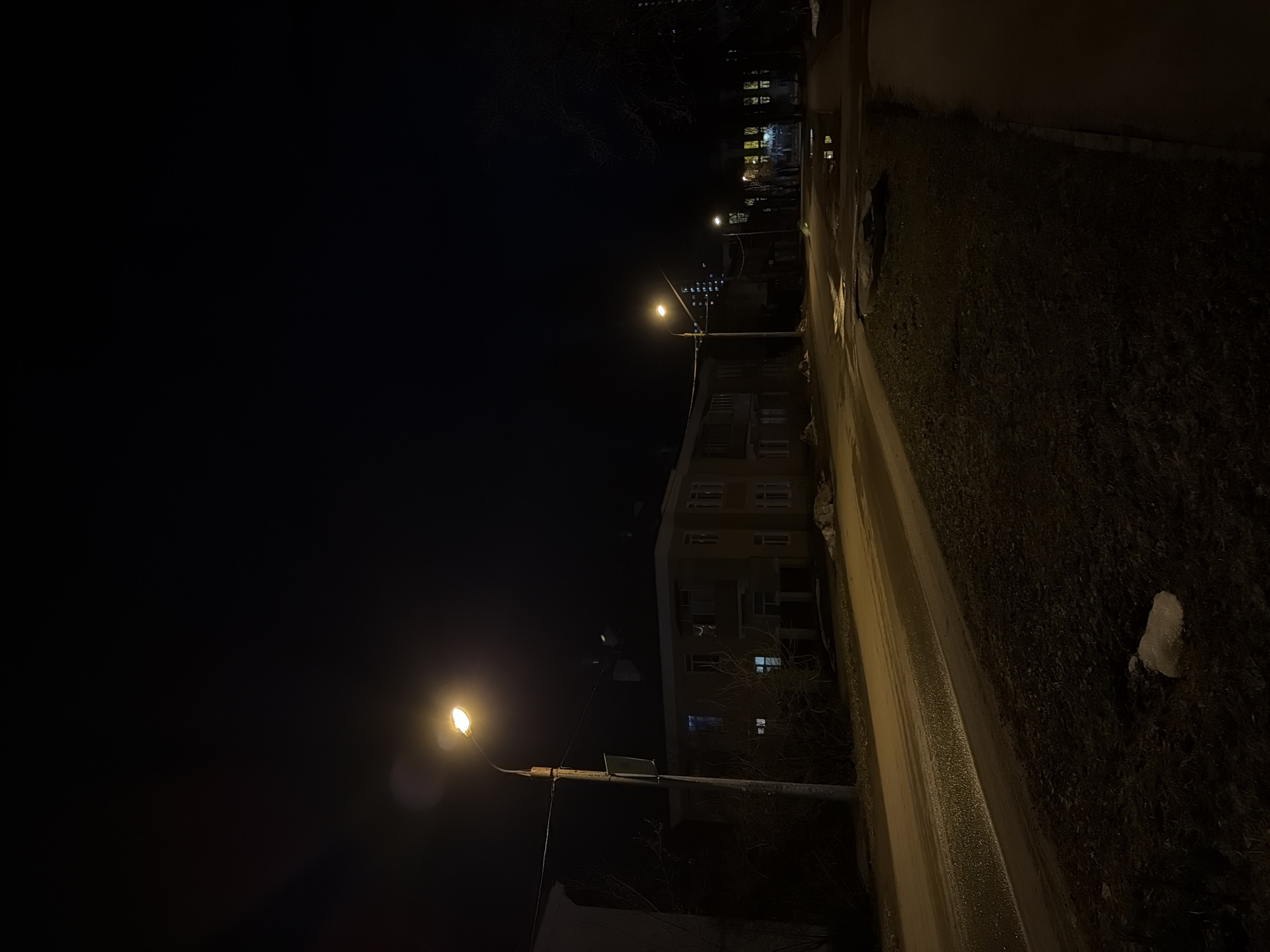 The image depicts a street scene at night in Perm, Russia. A wet road is illuminated by streetlights. Adjacent to the road is a dark, overgrown area. In the distance, there is a multi-story building with some lit windows. The sky is dark.