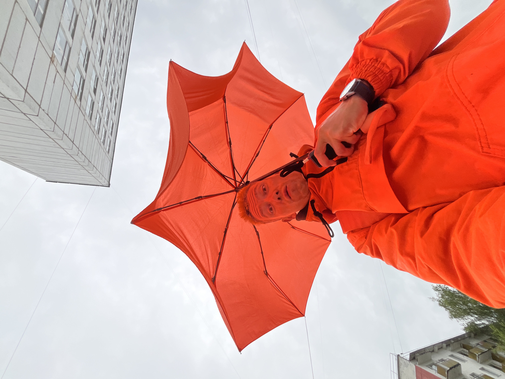 A man, viewed from a low angle, stands beneath a bright orange umbrella. He wears a matching orange jacket and has a surprised expression on his face. His left hand grips the umbrella handle, and a watch is visible on his right wrist. In the background, a modern, multi-story building with numerous windows looms against a cloudy sky. To the left, another building with balconies and a tree are partially visible. The scene appears to be set in an urban environment, possibly Chaikovsky, Russia, given the architectural style of the buildings and the overall atmosphere. The lighting suggests an overcast day.