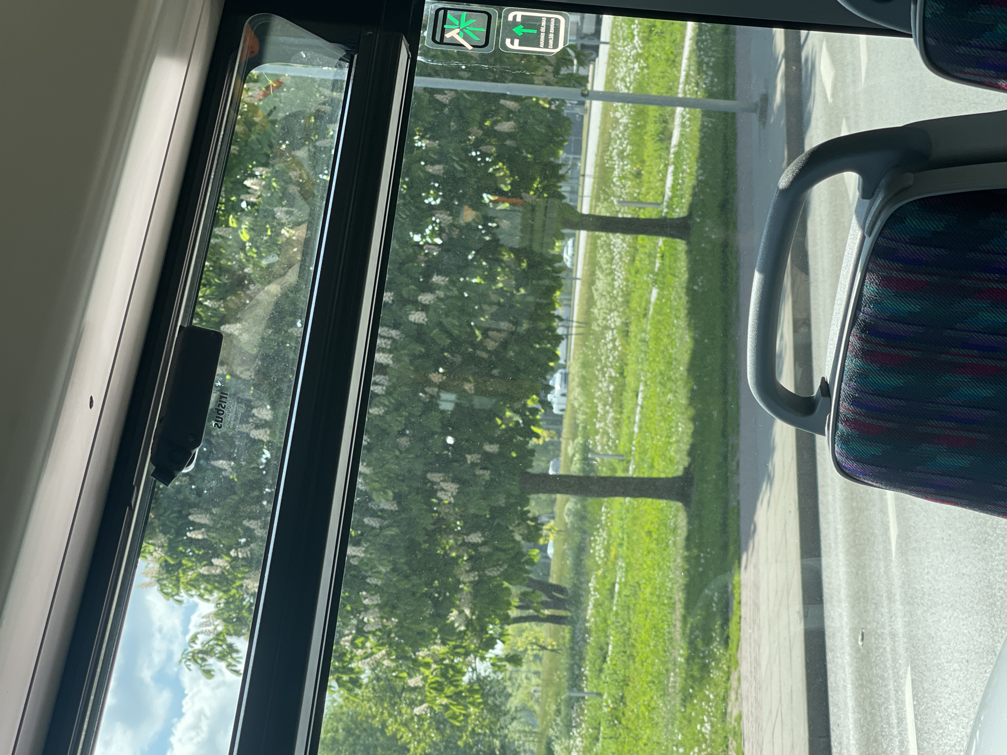A view from inside a vehicle, likely a bus, shows the outdoors through a window. The window frame is black and contains a safety hammer. Outside, there is lush green grass with white flowers, several trees, a lamp post, and a sidewalk adjacent to a road. A portion of a seat with a colorful patterned fabric is visible on the right side of the frame. The location is Unknown, Unknown.