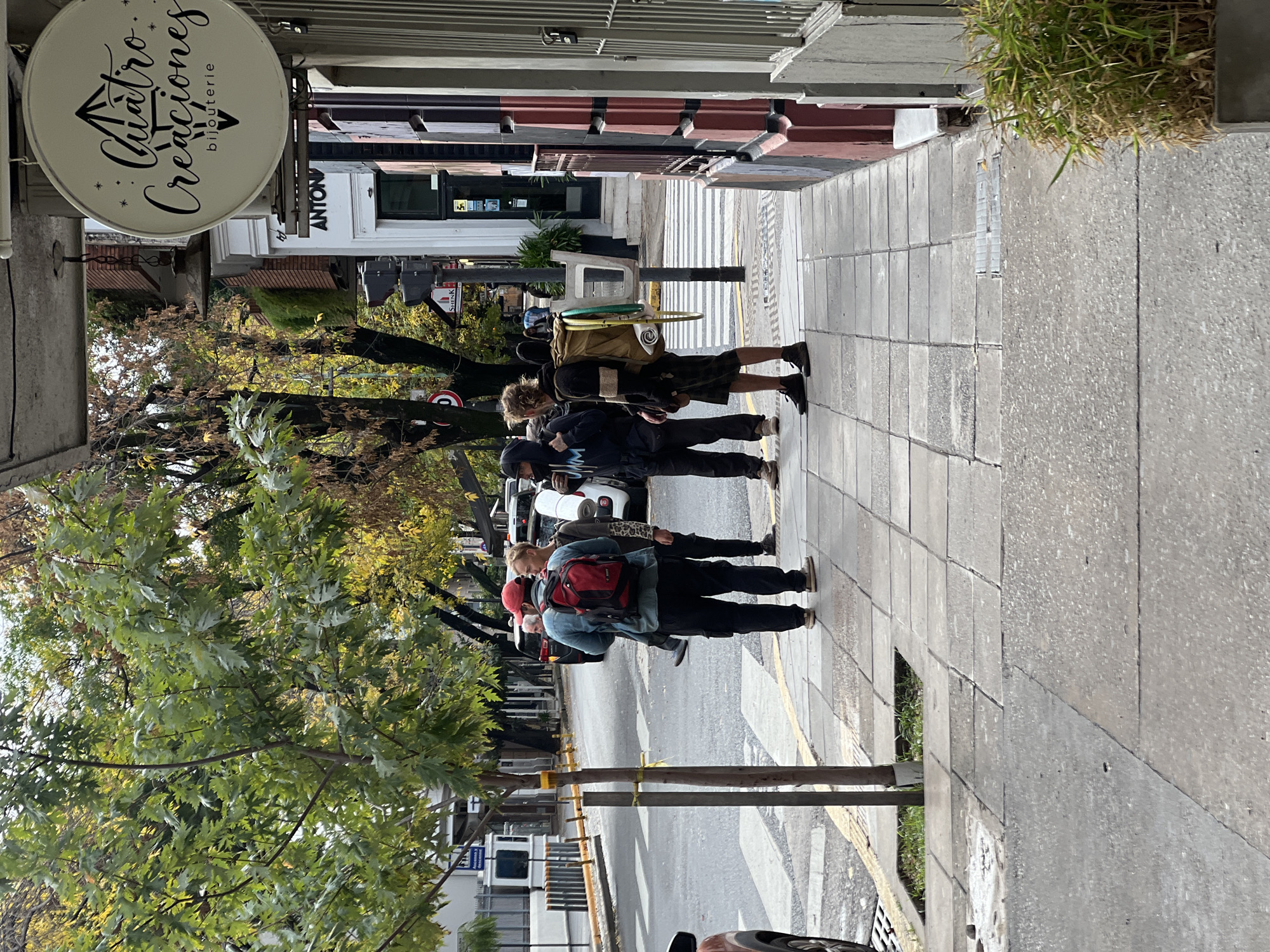 The image shows a street scene in Buenos Aires, Argentina. A group of five people are walking on the sidewalk. The first person wears a red hat and carries a red backpack. The remaining four are wearing dark clothing. Behind the group is a row of parked cars along a street with a crosswalk. A sign reads "Cuatro Creaciones bijouterie". Trees with green leaves and some yellowing leaves line the street. Buildings are visible in the background. The sidewalk is made of square concrete slabs.