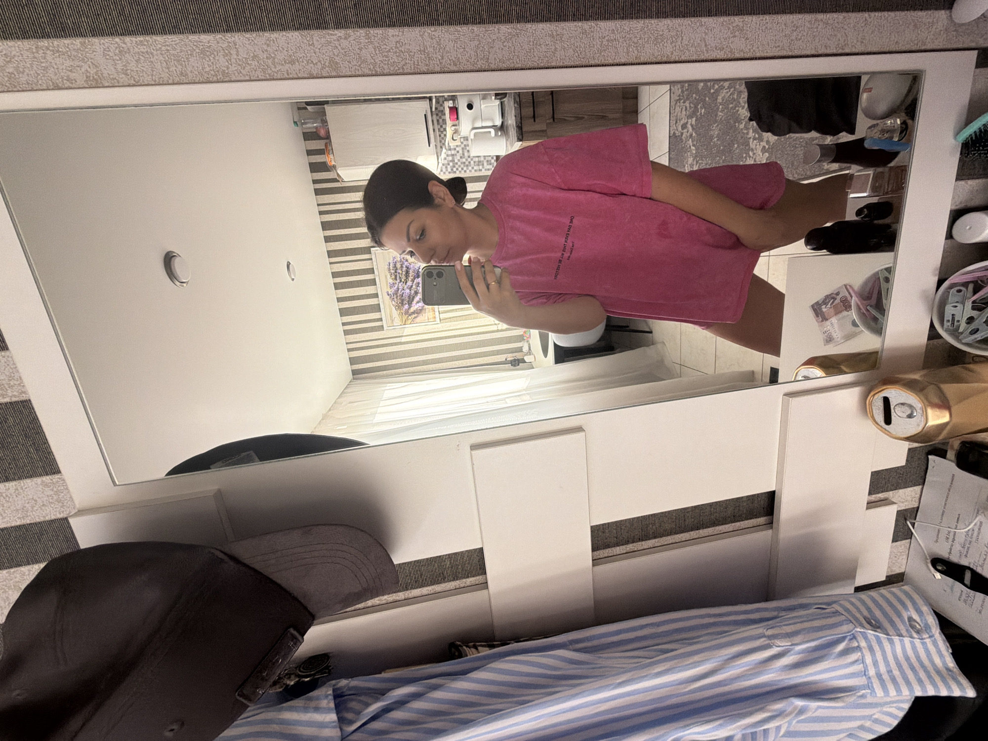 A young woman stands in front of a mirror, taking a selfie. She is wearing a pink t-shirt dress and has her hair pulled back in a bun. The mirror reflects a kitchen area with striped wallpaper. On a shelf next to the mirror, there is a grey baseball cap and a blue and white striped shirt hanging. The scene appears to be in an apartment.