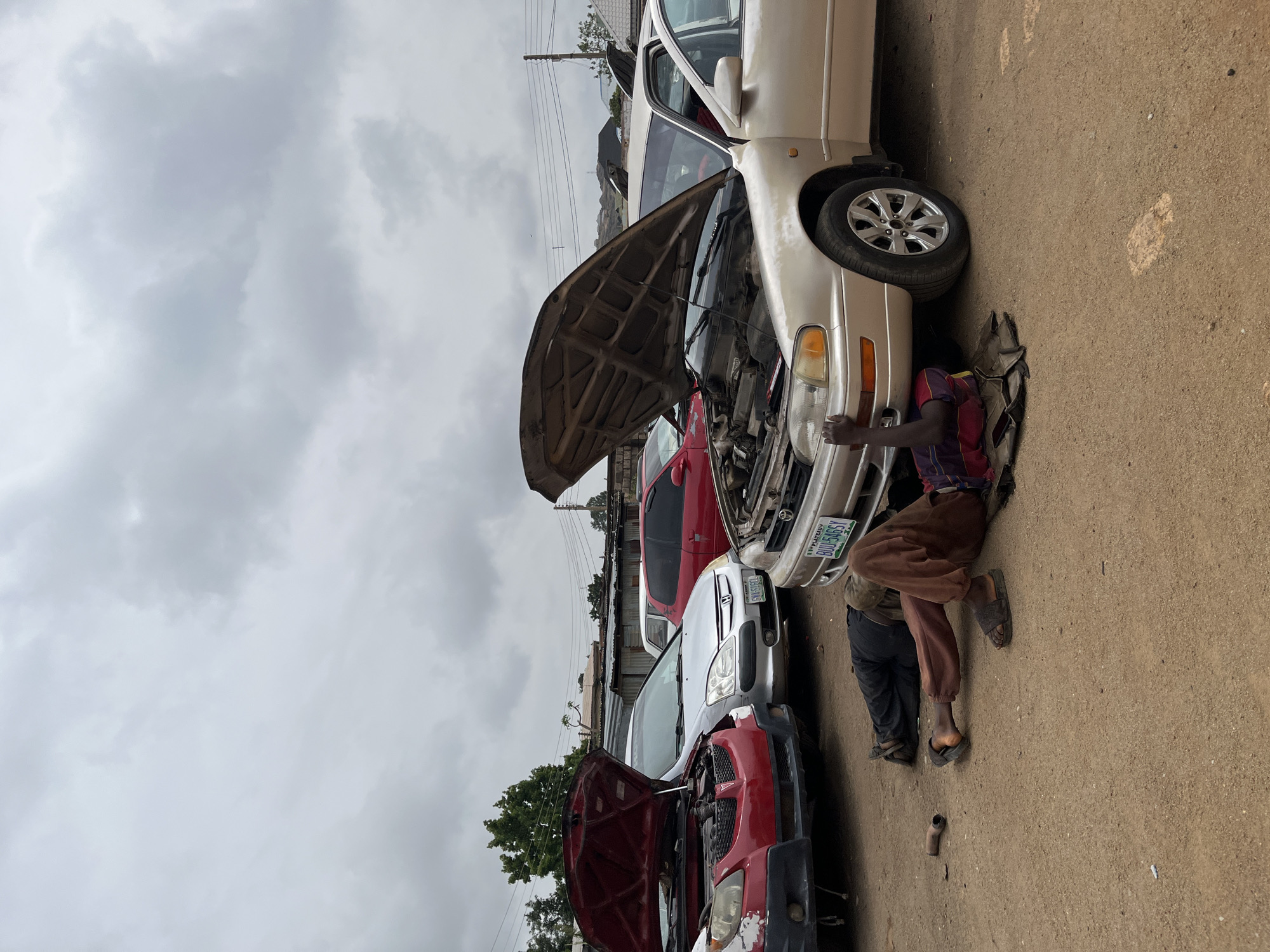 An outdoor scene captures an active auto repair area, likely in Jos, Nigeria, under an overcast sky. In the foreground, a mechanic is seen kneeling next to the front of a light-colored sedan, possibly beige or light gold, with its hood fully open, indicating work on the engine. The mechanic wears a multi-colored top and brown trousers, with sandals on his feet. Another person's lower legs and feet, clad in dark trousers and sandals, are visible further under the same car, suggesting they are also working or assisting.
The sedan features chrome-finished wheels and has a Nigerian license plate that reads "BUE 546GY". Tools or car parts are scattered on the dirt ground near the workers. Behind this car, several other vehicles are parked in various states of repair or waiting. A red car directly behind the sedan also has its hood open. Further in the background, a silver/grey car and another red car are visible, parked along a row of corrugated metal fences or structures. Overhead power lines and distant trees complete the cluttered, industrial-like setting. The overall impression is one of active, informal vehicle maintenance taking place during the daytime. Another visible license plate on a car in the middle background appears to be "SM3 686".