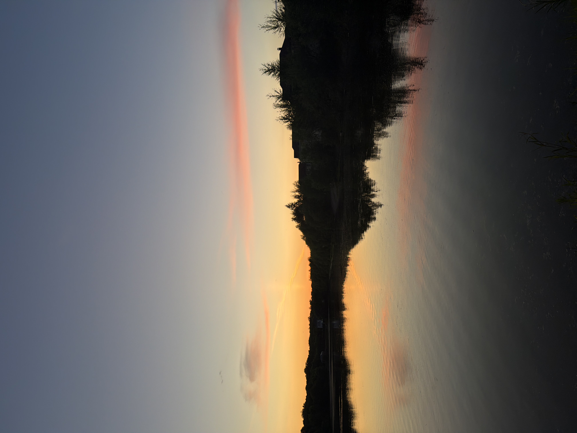A serene sunset over a tranquil body of water. The sky displays a gradient of soft blues transitioning to warm oranges and pinks near the horizon, with wispy clouds scattered across the upper atmosphere. A silhouetted treeline forms the backdrop, with several houses and scattered trees visible amongst the foliage. The water's surface mirrors the sky and landscape, creating a peaceful reflection. The scene suggests a quiet evening in a natural or semi-rural setting.