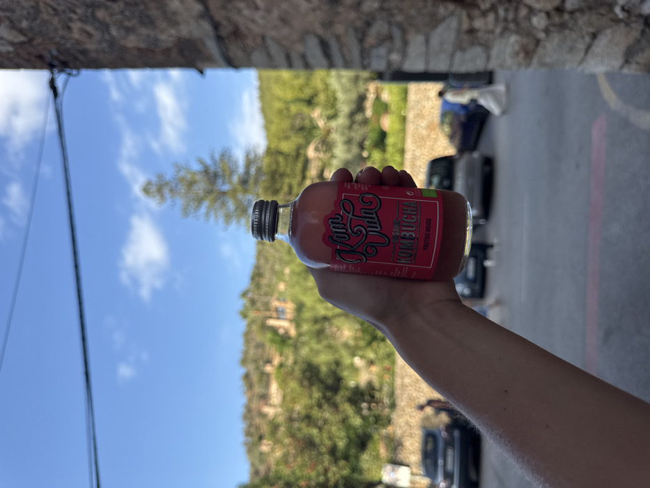 A close-up, eye-level shot captures a light-skinned human hand holding a clear glass bottle of "Kom Vida Organic Kombucha - Frutos Rojos" in the foreground, against a blurred backdrop of a street scene in Deià, Spain. The bottle contains a reddish-pink liquid and has a metallic screw cap. In the midground, a paved road or parking area is visible with several blurred vehicles, including a dark car on the left and a blue car on the right. Two indistinct figures are present on the street: one near the dark car on the left, and another, clad in light-colored clothing, walking from right to left in the distance. The background features lush, green, tree-covered hills characteristic of Deià, with several traditional buildings possessing reddish roofs nestled among the foliage. A prominent tall conifer tree stands out against the clear blue sky, which has sparse white clouds. The scene is illuminated by natural daylight.