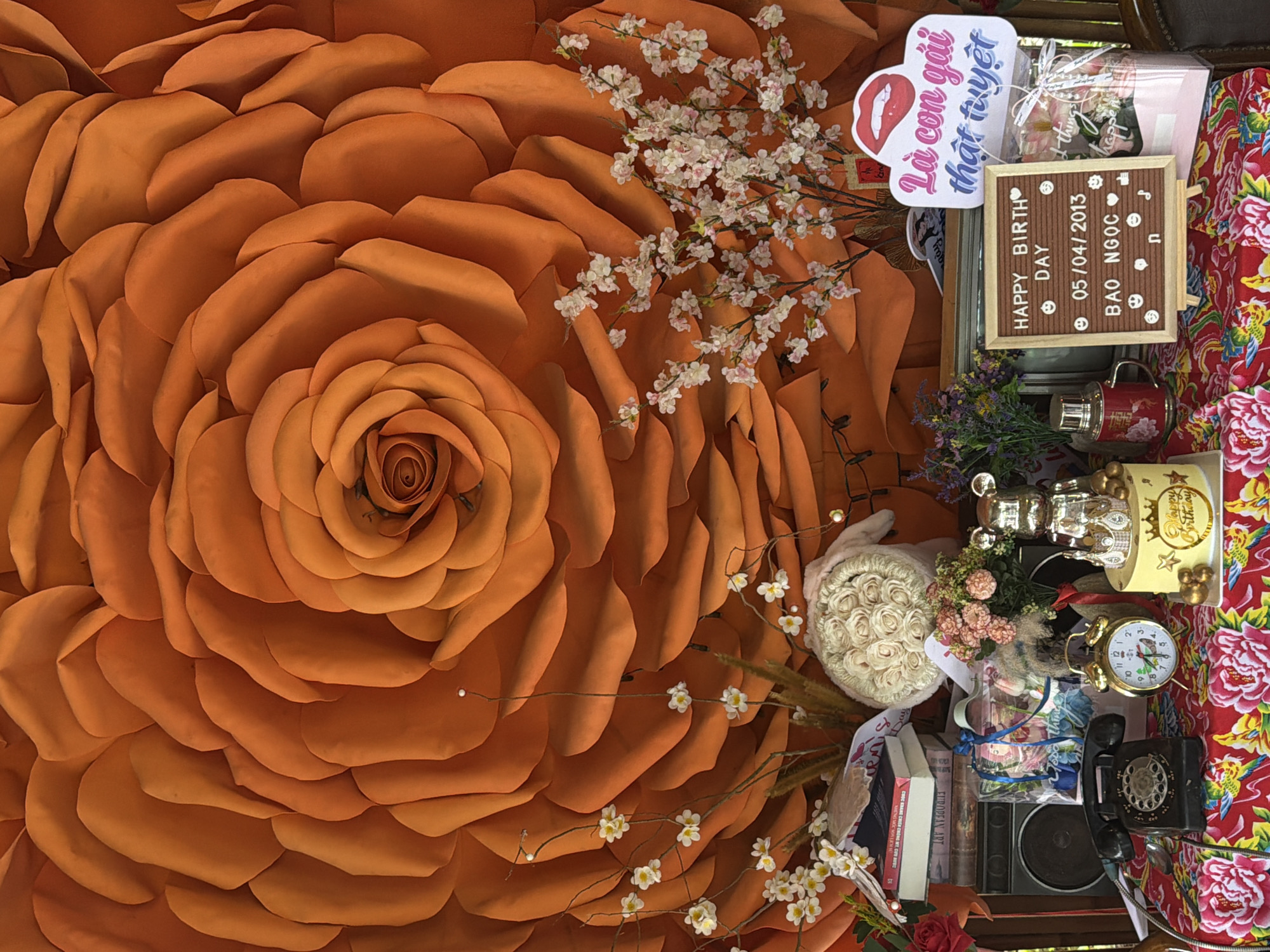 Here's a description of the image:
The image is a close-up of a decorative display, dominated by a large, orange, artificial rose. Sprigs of white flowers are arranged around the right side of the rose. Various objects are clustered together on what appears to be a table or shelf, covered with a floral-patterned fabric.
Objects include a small message board with "HAPPY BIRTHDAY DAY 05/04/2013 BAO NGỌC" displayed. A speech bubble with Vietnamese text ("Là con gái thật tuyệt") is also present. Other items include a small floral gift, a vase, a vintage-style alarm clock, a vintage telephone, a radio speaker, books with text partially visible like "EUROPEAN ART" and "NHỮNG MỐC SON LỊCH S", and various decorative trinkets including a ceramic rabbit.
The scene suggests a staged backdrop or photo booth setup. Without further information, pinpointing a specific location in Đà Nẵng, Vietnam, is not possible.