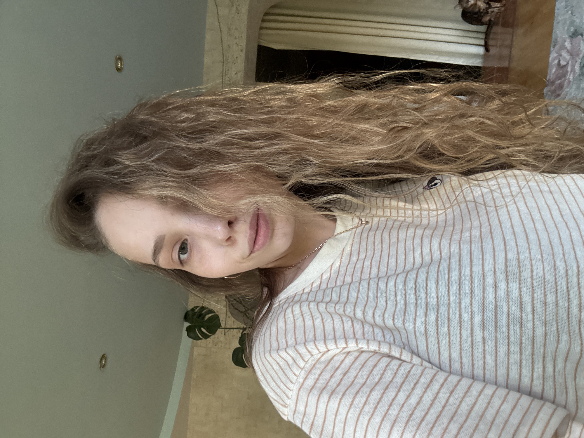 A young woman with long, wavy blonde hair is pictured in a medium shot, facing left and looking slightly away from the camera. She is wearing a striped shirt with thin, light brown stripes on a cream background. A delicate gold necklace is visible around her neck. Her skin is fair, and she has light-colored eyes. Her mouth is slightly parted, revealing a natural expression.
The background suggests an indoor setting, likely a home. The wall behind her is painted a light, neutral color, and a circular light fixture is visible on the ceiling. To the right, a doorway or alcove is visible with drawn blinds, and part of a patterned curtain or tapestry is seen. A large houseplant with distinctive leaves, possibly a Monstera, is situated in the lower left of the frame, adding a touch of nature to the interior.
The lighting appears to be natural, suggesting it might be daytime, possibly late afternoon given the warm tones. The overall mood is relaxed and casual. There is no visible text in the image. The setting, based on the provided location context of "Прикамский, Russia," places the scene within this region.